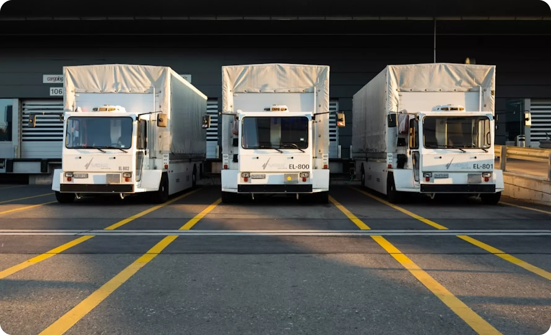 Three white airport cargo vehicles parked side by side in a lot with yellow parking lines, in front of a dark building with vents.