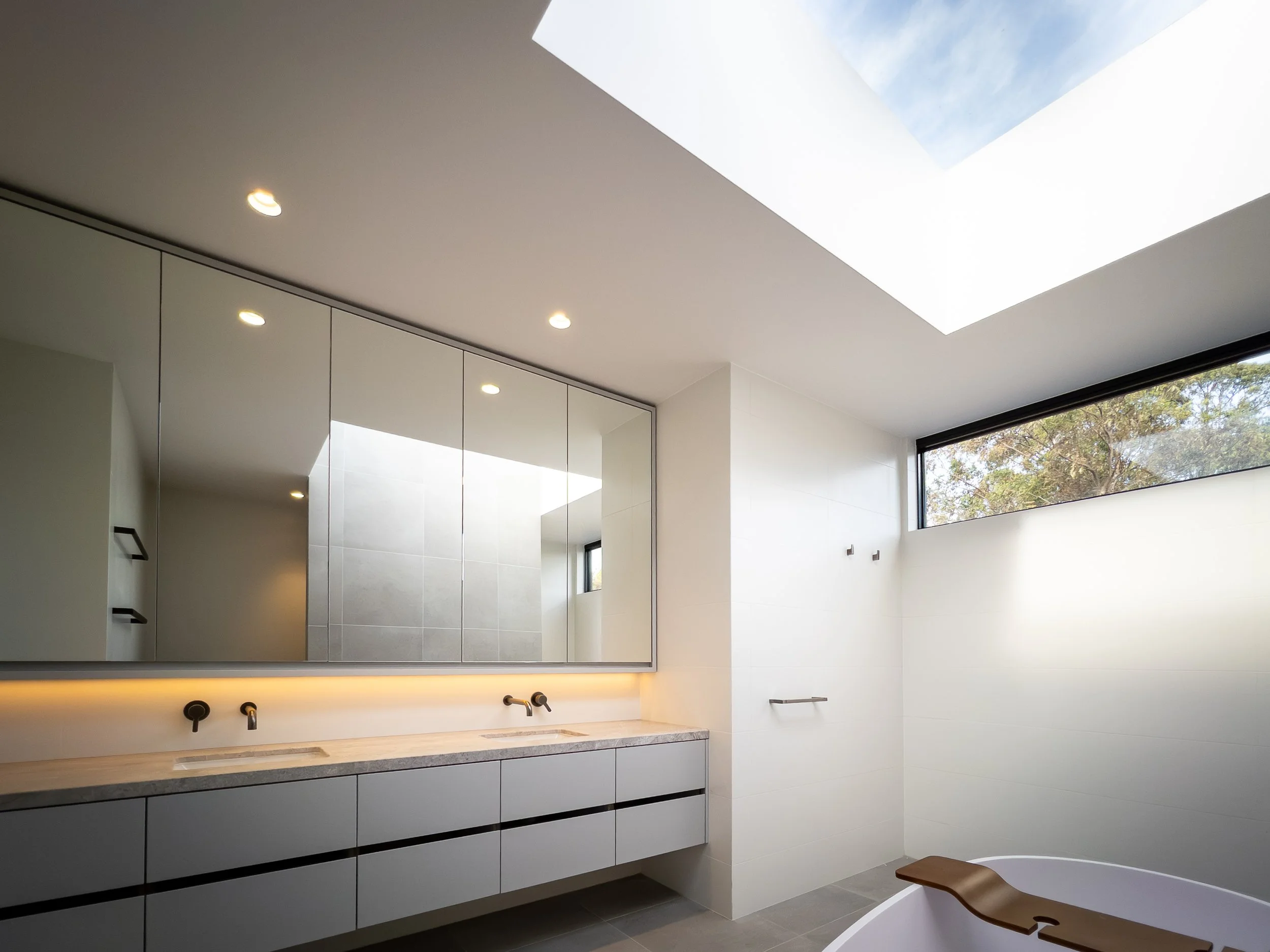 Modern minimalist bathroom with a large mirror, white cabinets, double sinks, a bathtub with a wooden tray, and a long horizontal window near the ceiling showing trees outside, under a skylight in a bright room.
