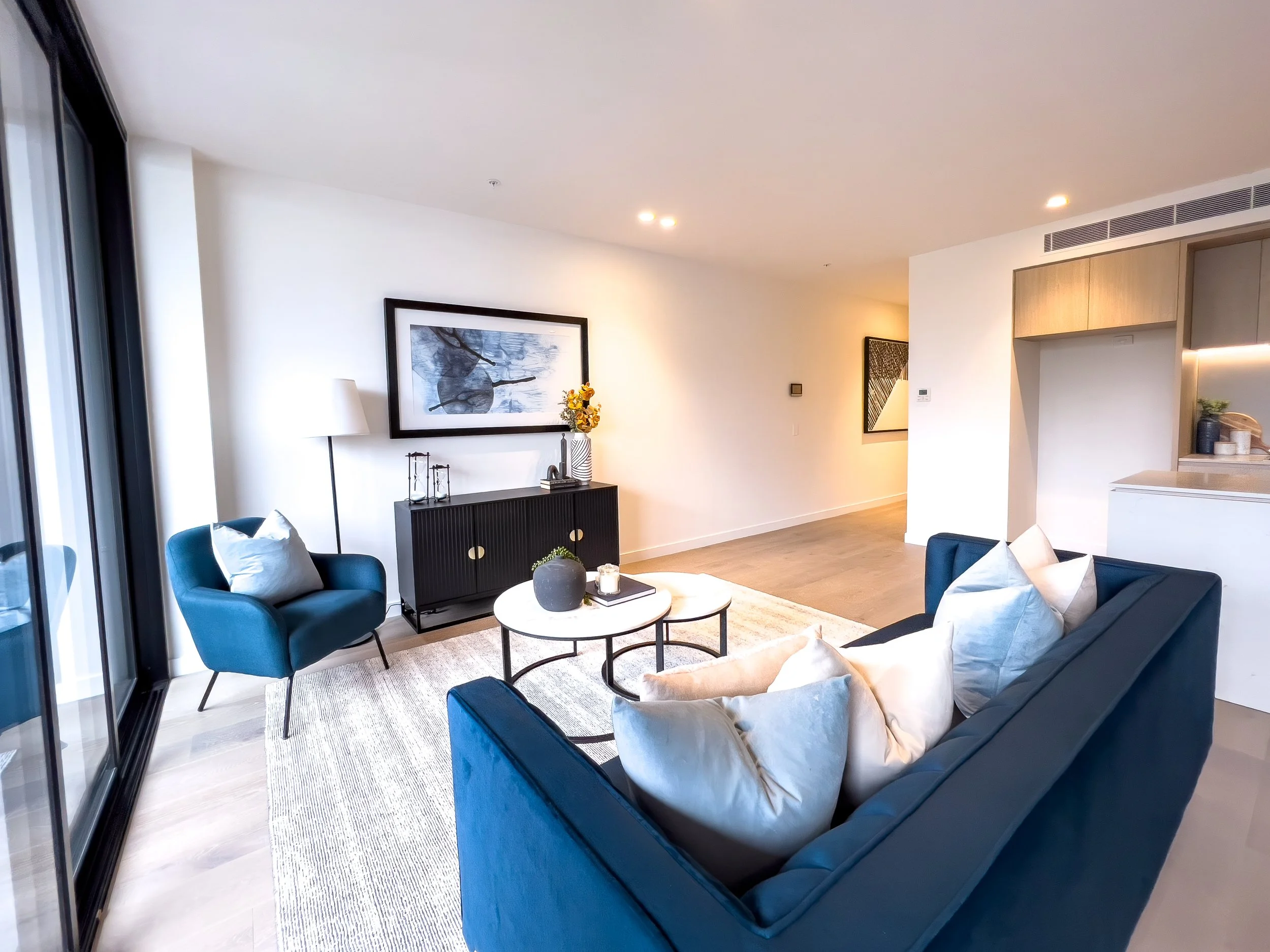 Living room with navy blue sofa, matching armchair, white pillows, black sideboard with artwork and flowers, two white round coffee tables, large glass sliding doors, framed artwork on walls, wooden floors, and minimal decor.