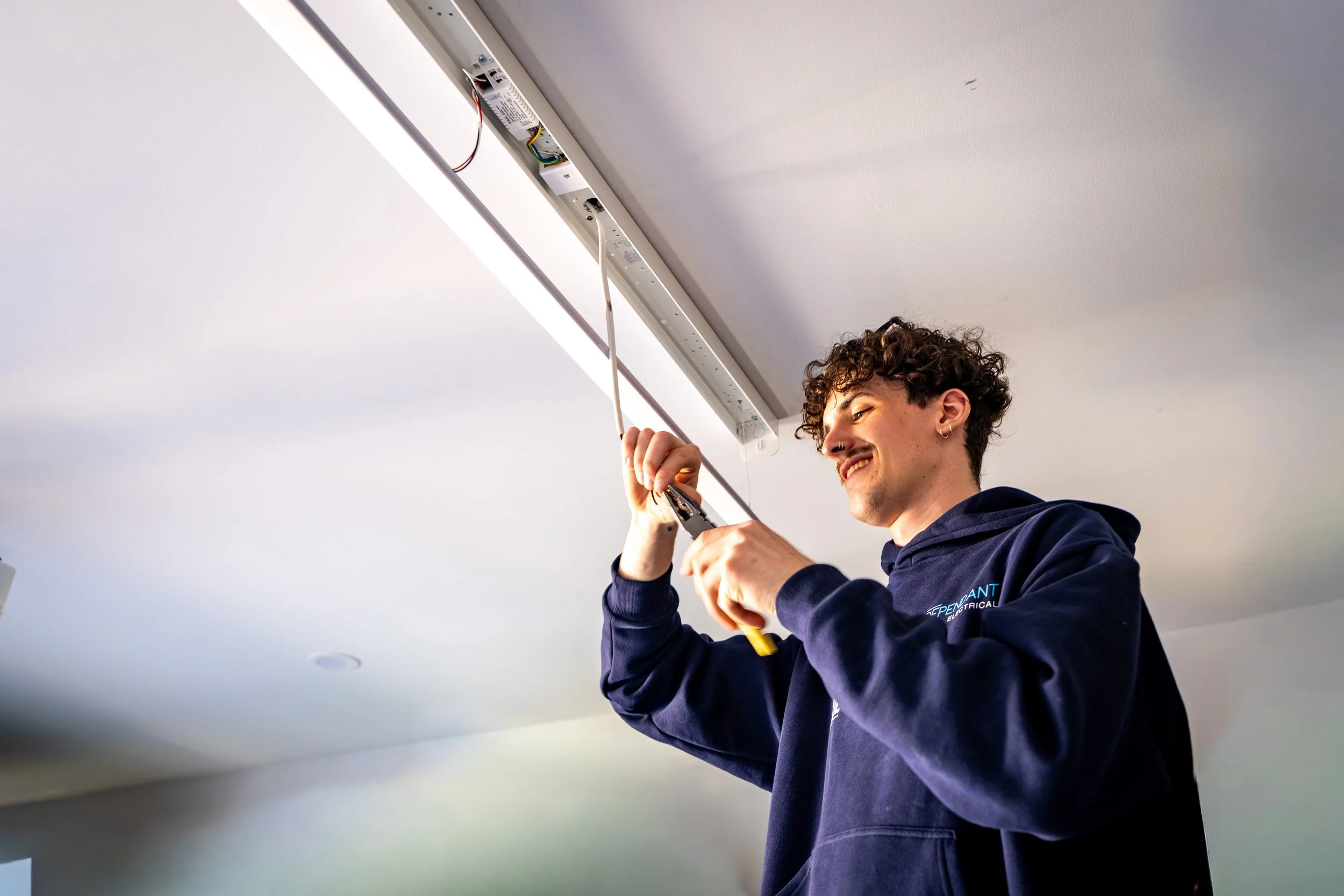 A young man is installing or repairing a ceiling light fixture with a screwdriver in a room with a white ceiling and wall.