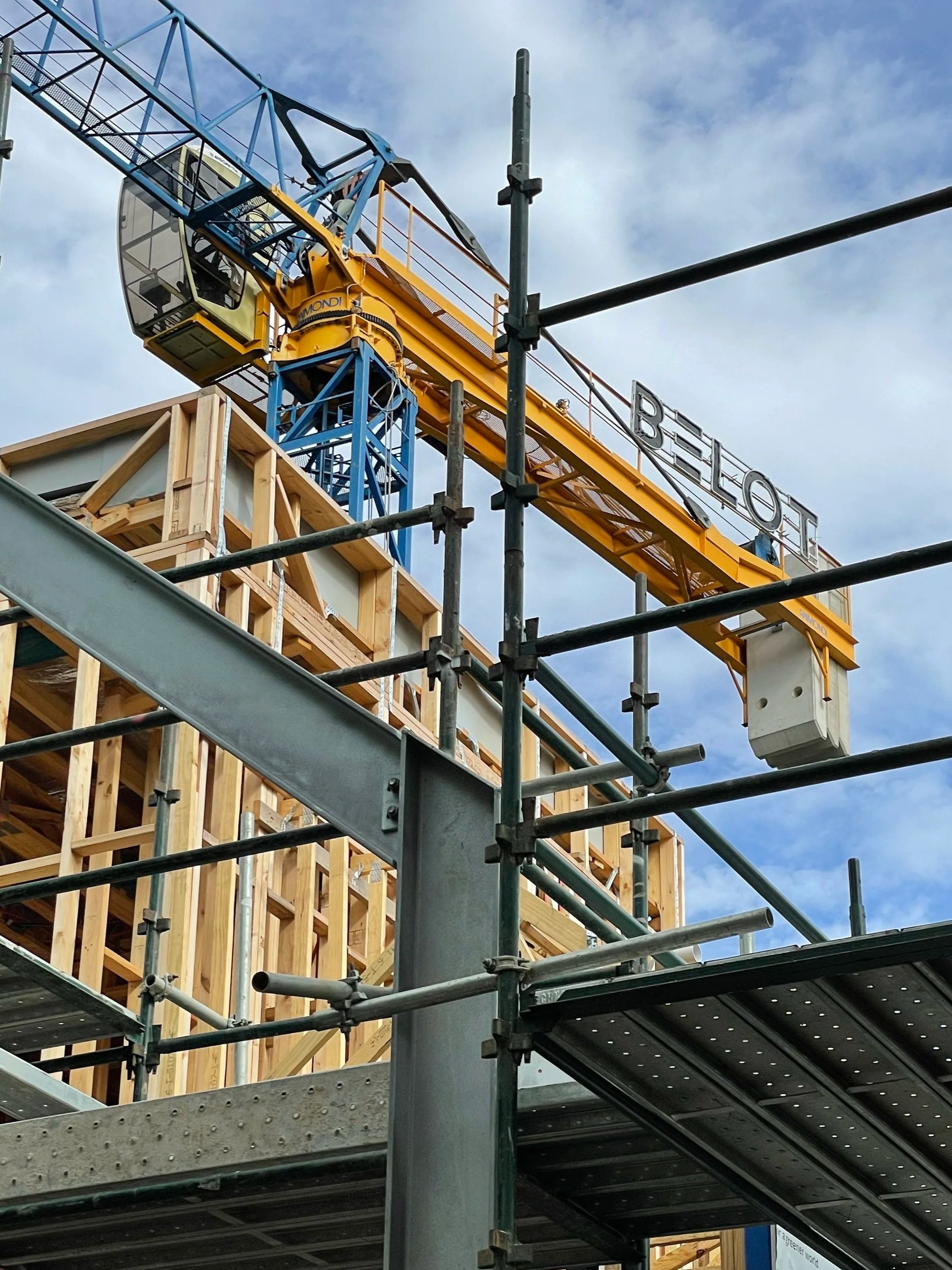 Construction site with a yellow and blue tower crane and a wooden building frame surrounded by metal scaffolding under a partly cloudy sky.