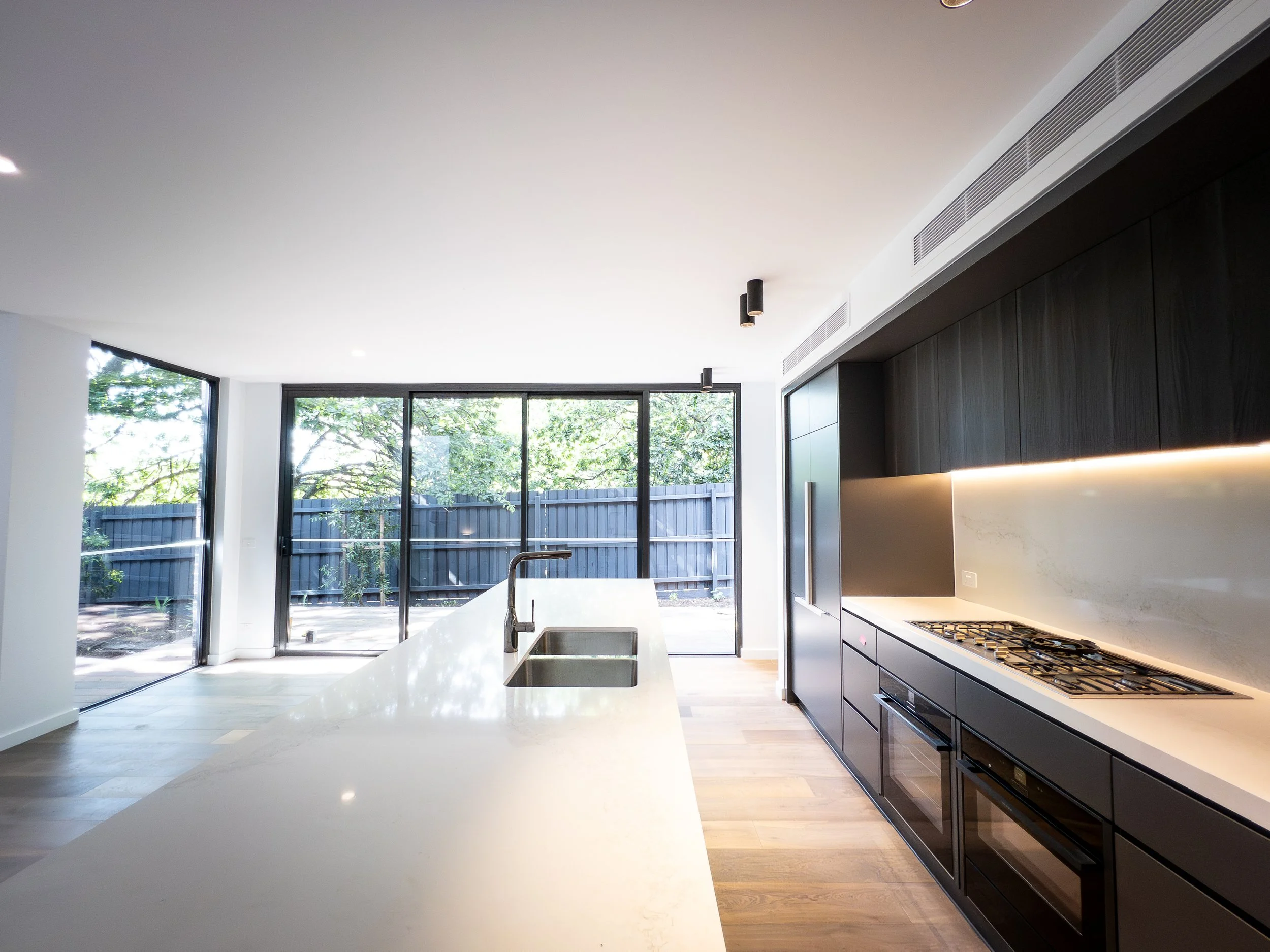 Modern kitchen with white island, black cabinetry, gas stove, and large windows overlooking a fenced yard with trees.