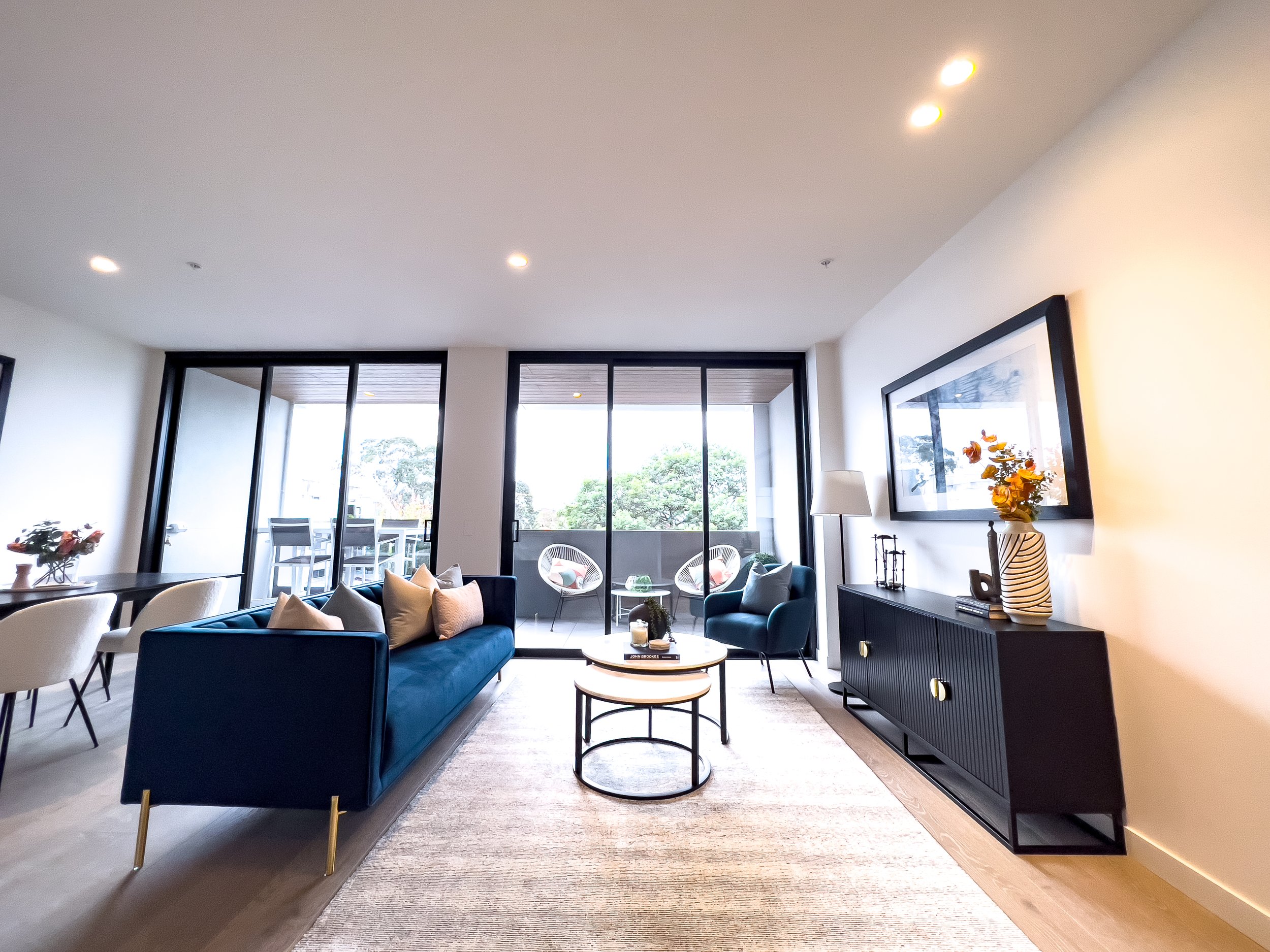 Modern living room with a blue sofa, armchair, coffee table, black sideboard, and sliding glass doors leading to a balcony with outdoor seating, decorated with artwork, vases, and cushions.