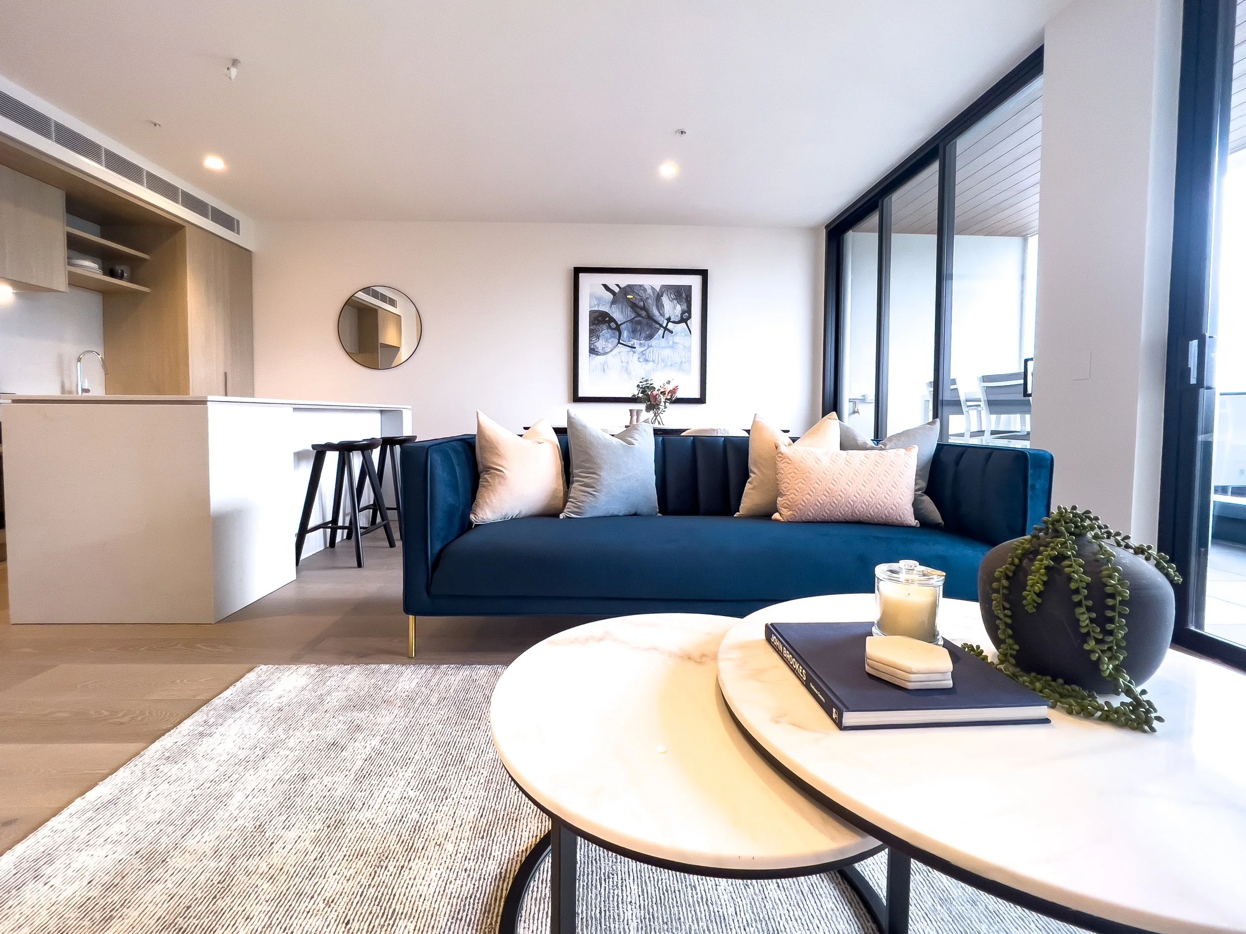 Modern living room with a blue velvet sofa, white and gray pillows, round coffee tables with a candle, book, and plant, large sliding glass doors leading to a balcony, and artwork on the wall.