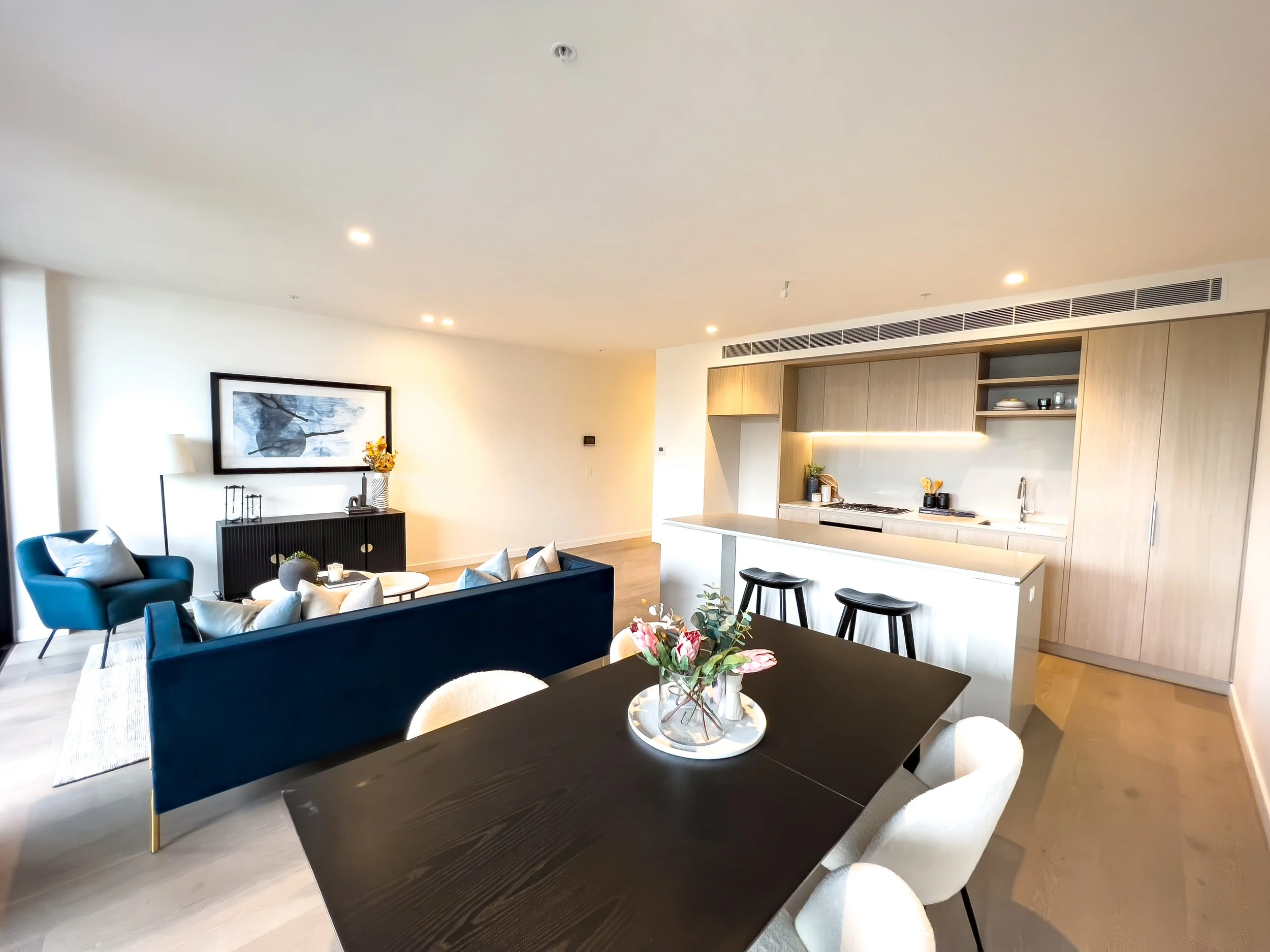Open-concept living and dining area with a white kitchen, a blue sofa, armchair, coffee table, framed artwork, and a dining table with beige chairs, decorated with flowers.