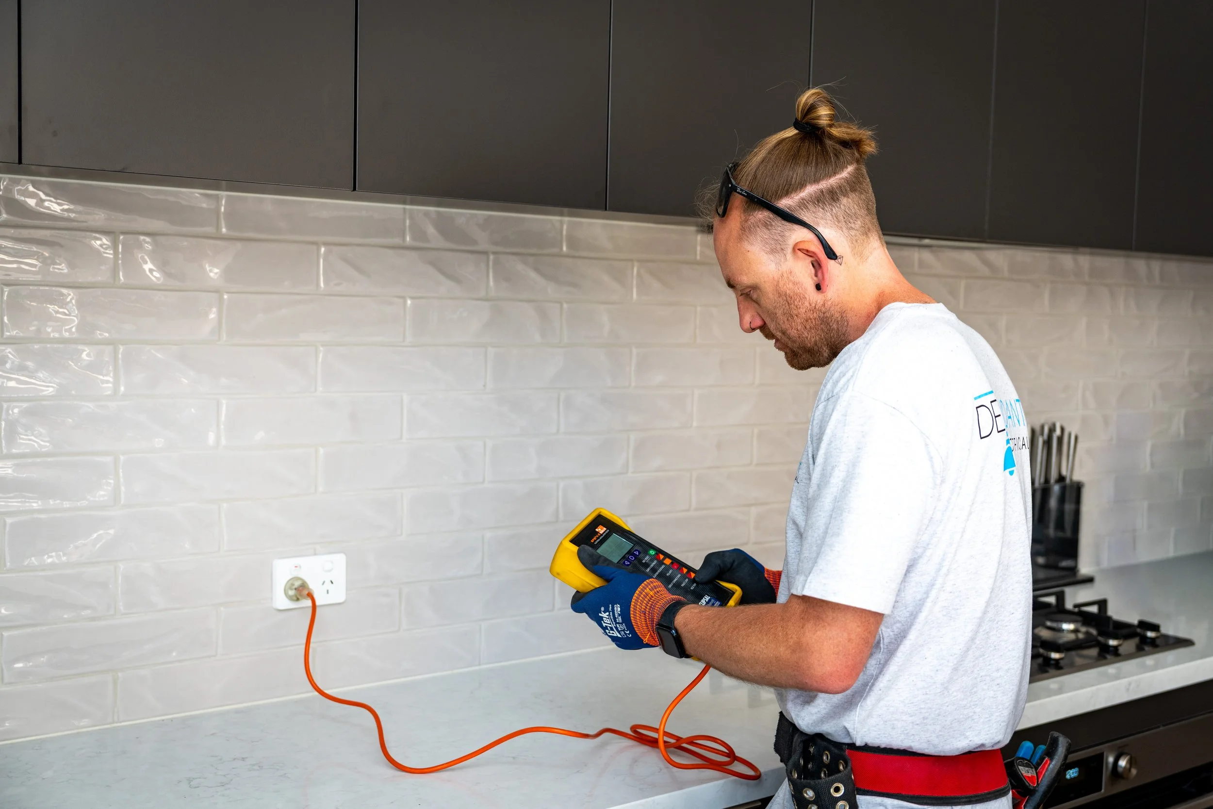 A technician wearing gloves and a tool belt checks electrical wiring with a multimeter in a modern kitchen.