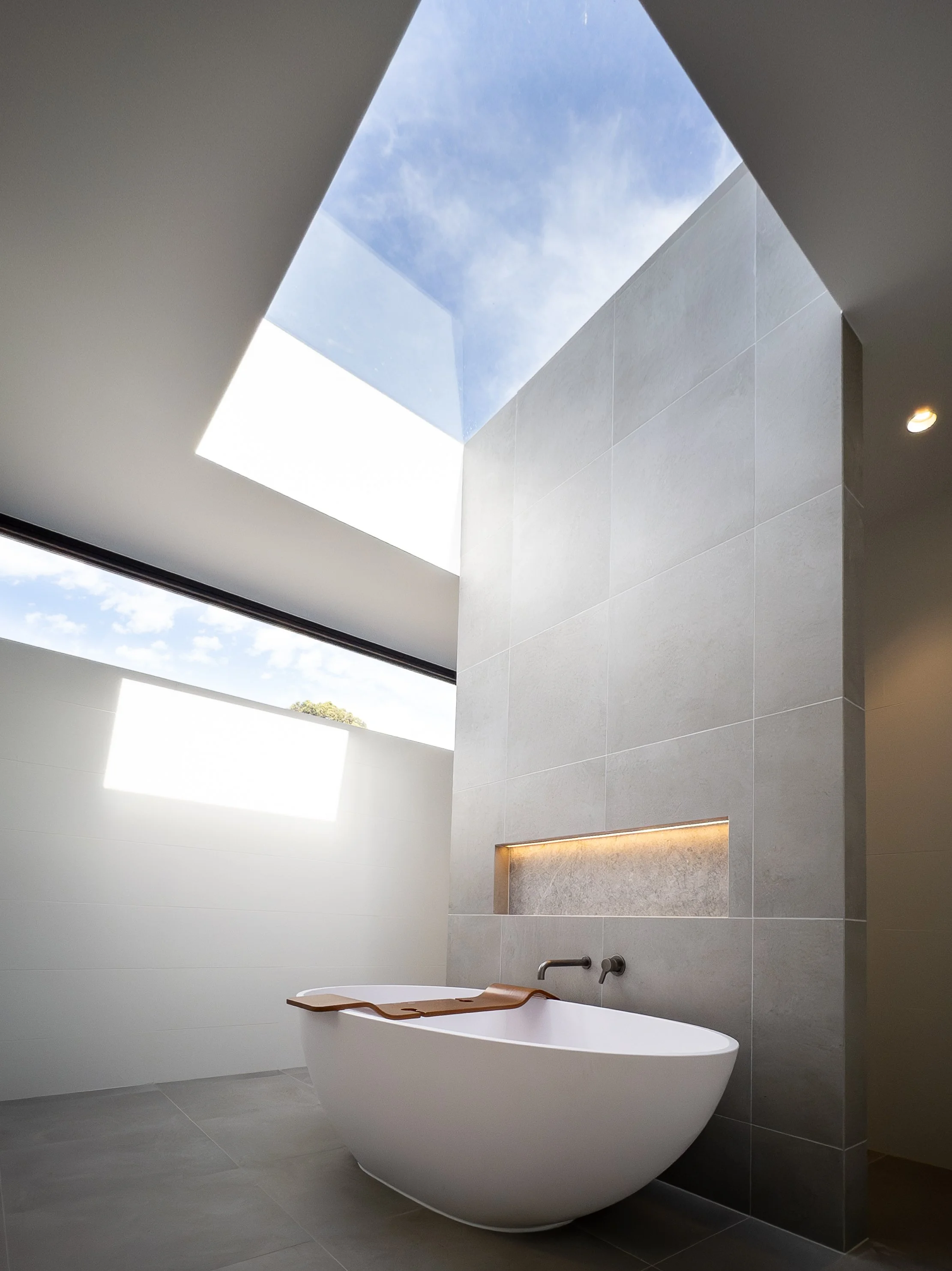 Modern bathroom with large skylight, freestanding bathtub, and minimalist gray tiles.