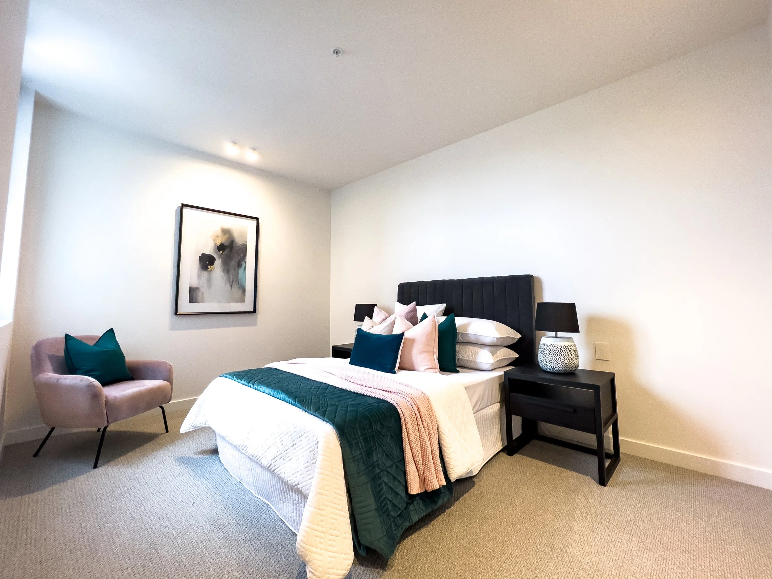 A modern bedroom with a bed, black headboard, multiple pillows, a side table with a black lamp, a pink armchair with a teal pillow, and a framed abstract artwork on the wall.
