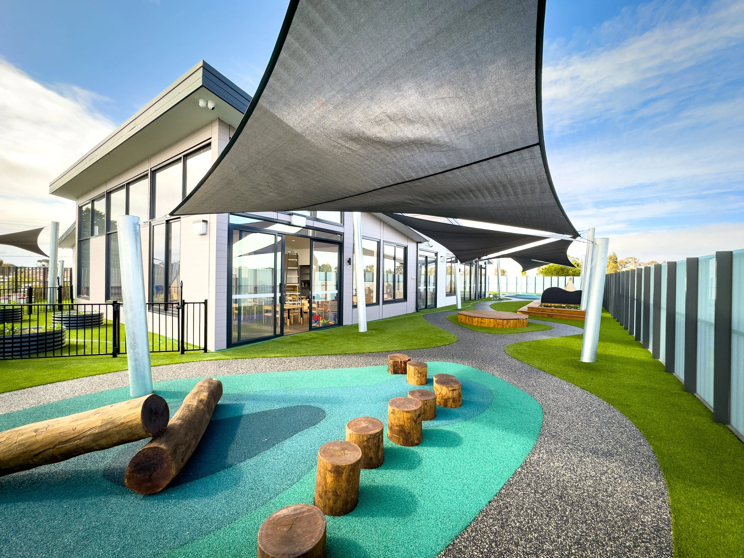 Outdoor playground area with artificial grass, shaded canopy, logs, and a winding gravel path adjacent to a modern building with large glass windows and a fenced yard.