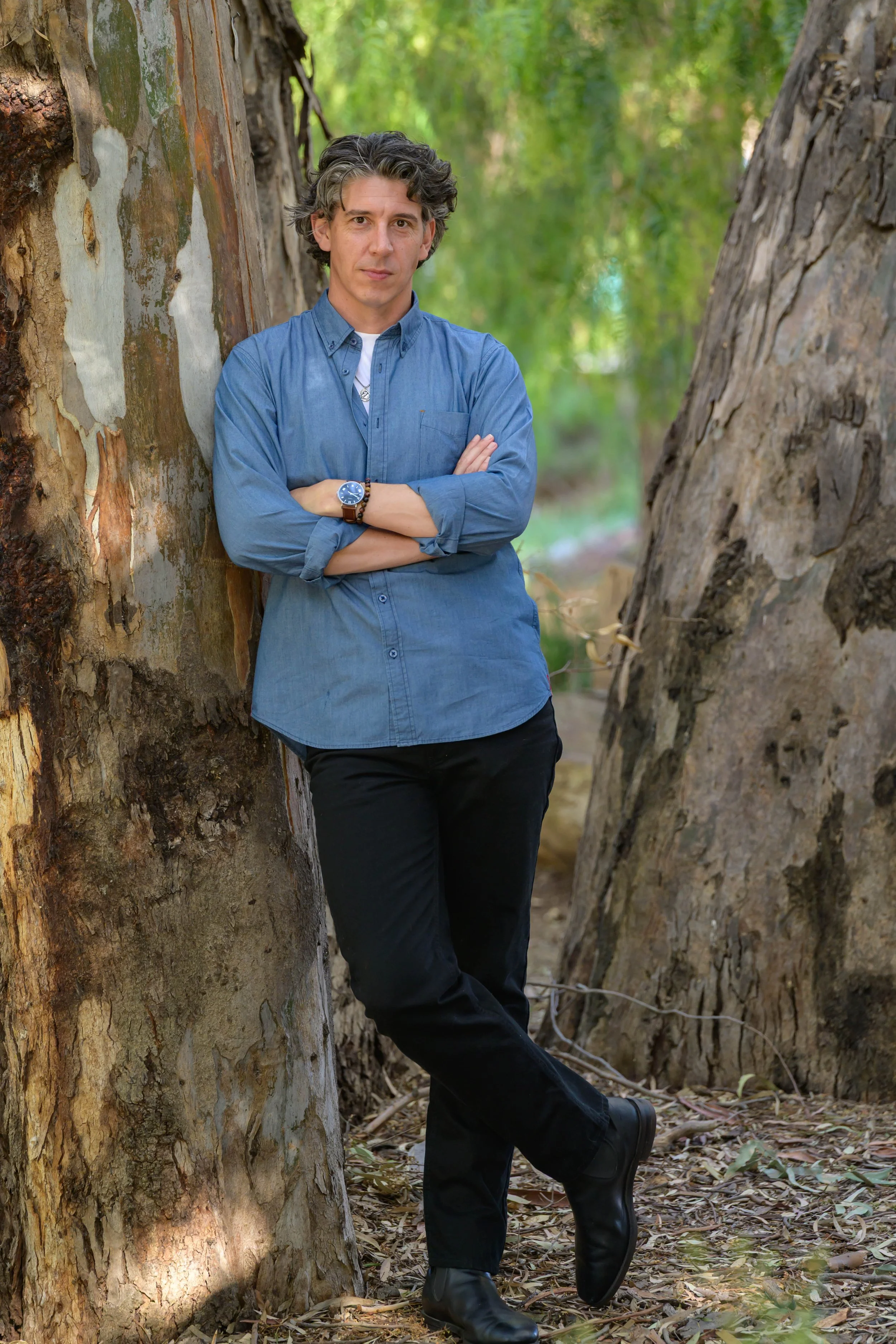 A man standing outdoors, leaning against a large tree with arms crossed, surrounded by green foliage
