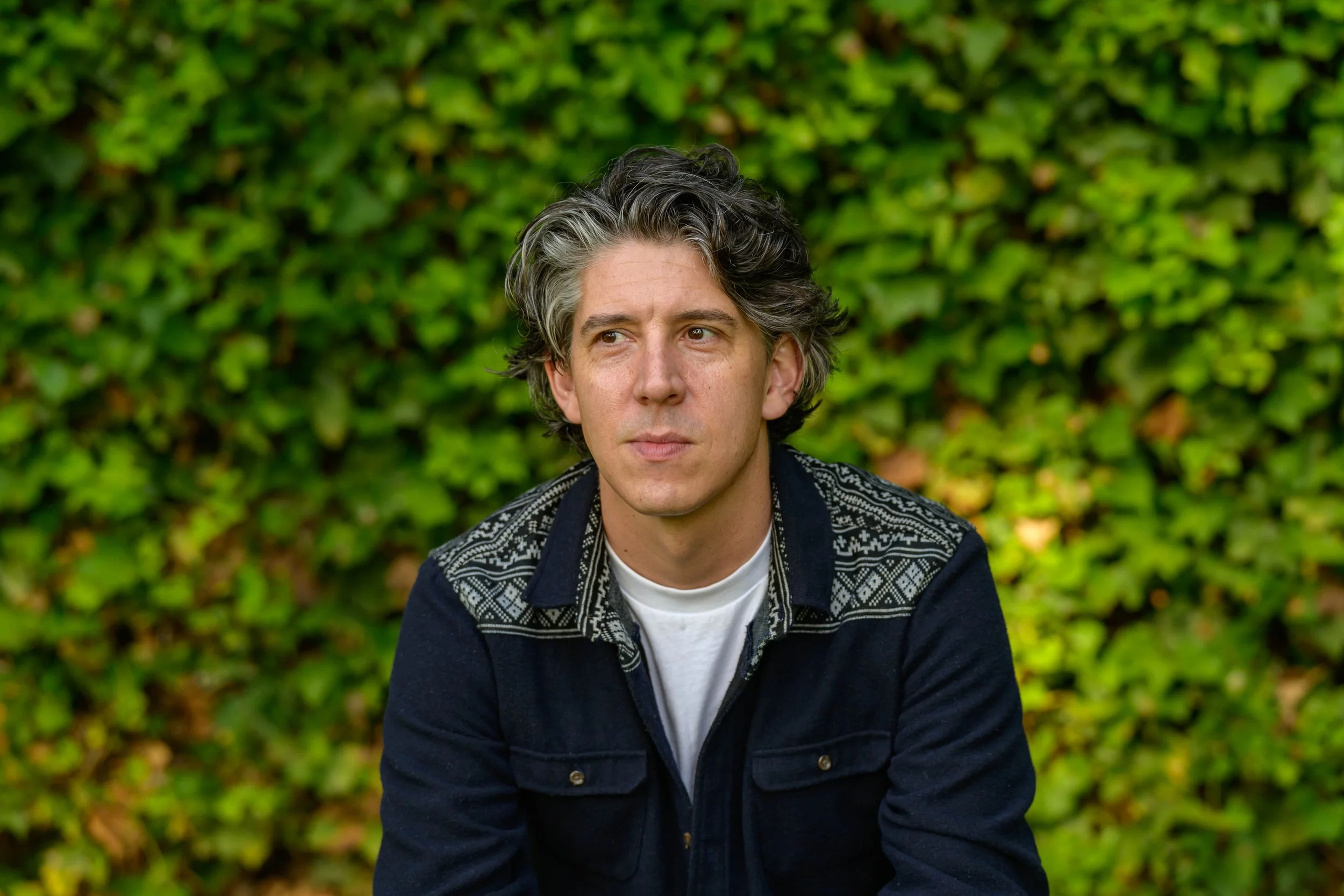 A man with gray and black hair sits outdoors in front of a lush green hedge, wearing a dark jacket with intricate patterns on the shoulders.