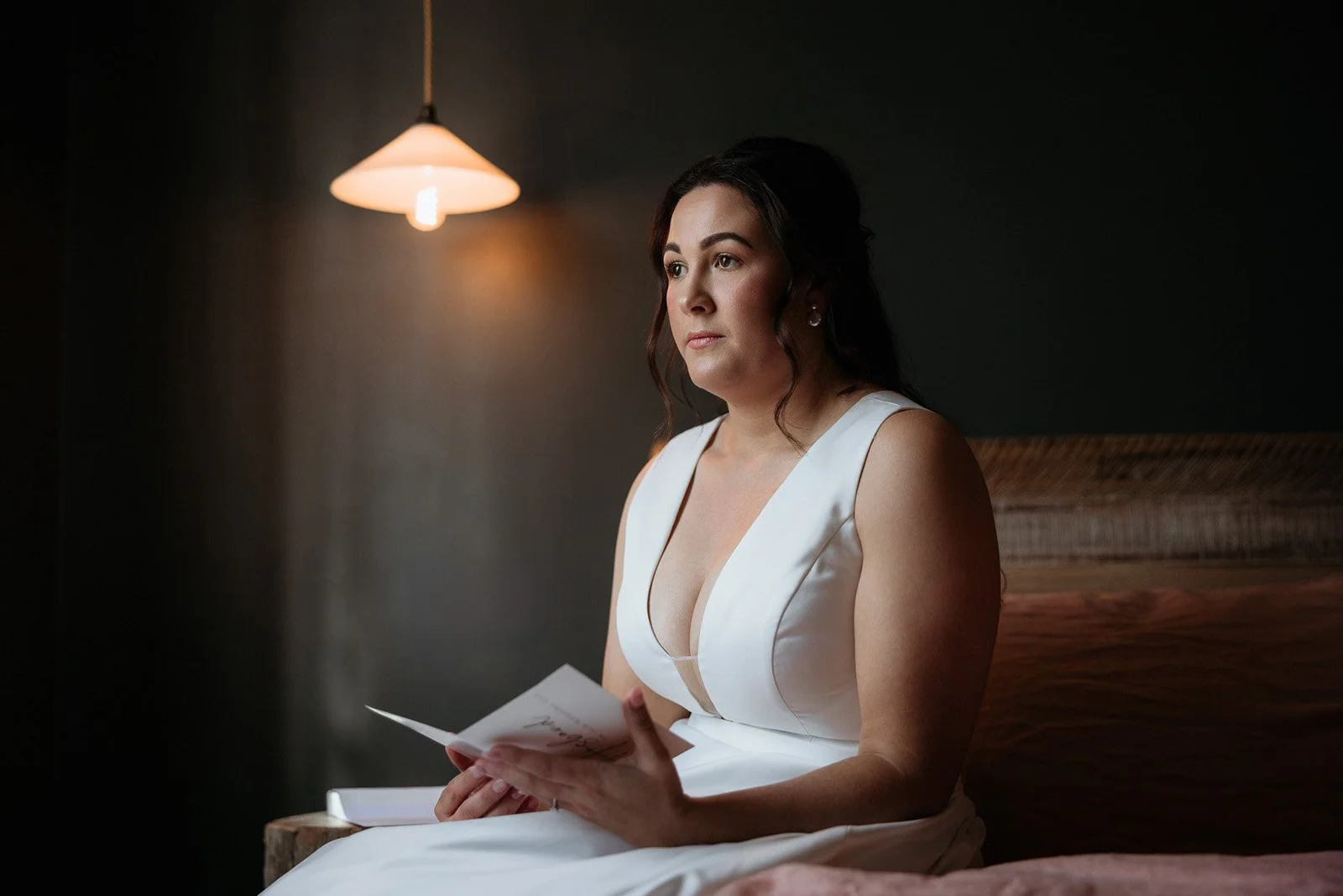 Woman in white dress sitting on bed, holding a piece of paper, in a dimly lit room with a hanging lamp.