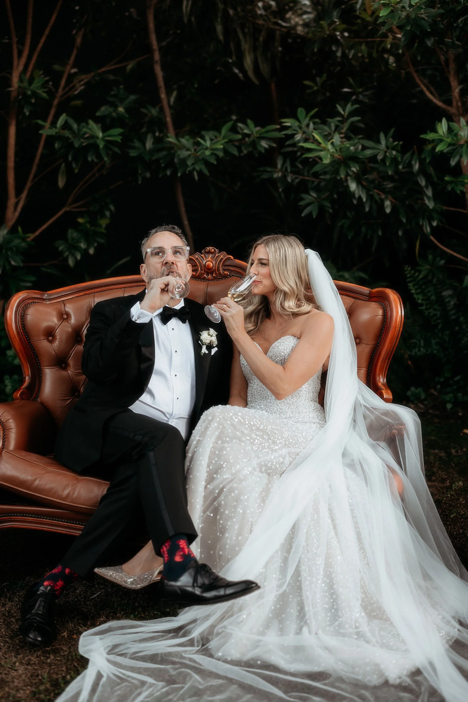 A bride and groom sitting on a vintage leather sofa outdoors, both drinking champagne. The groom is wearing a black tuxedo with a bow tie, glasses, and casual shoes. The bride is in a white wedding gown with 3D textured beading, a veil, and silver shoes. They are surrounded by lush green trees.