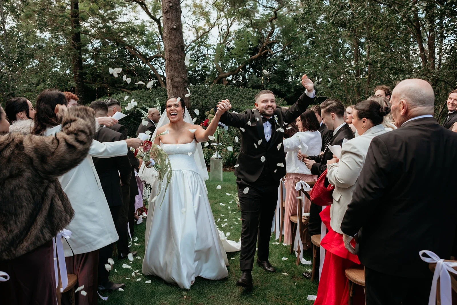 A bride and groom walking hand in hand through a wedding aisle, smiling and celebrating surrounded by guests throwing flower petals outdoors in a lush green garden.