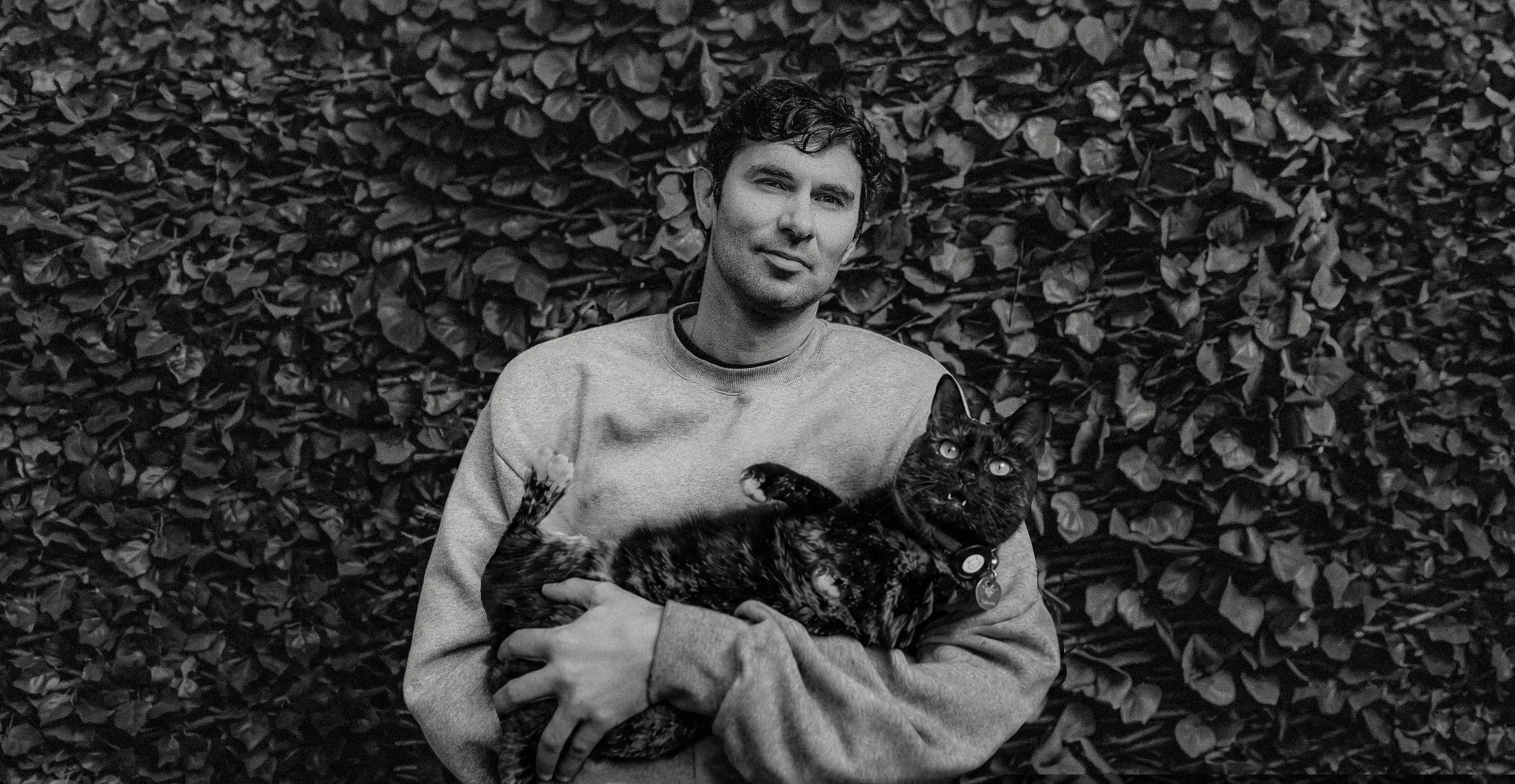 A young man with curly hair and a light-colored sweatshirt holding a black cat with a collar in front of a dense hedge.