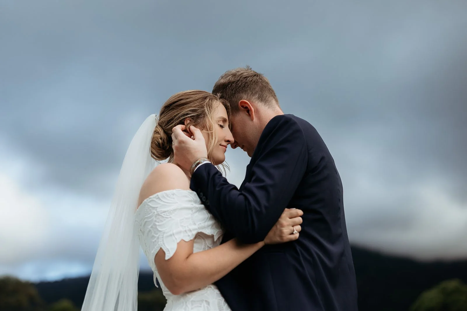 A bride and groom sharing a romantic moment outdoors, with their foreheads touching and eyes closed, under a cloudy sky.