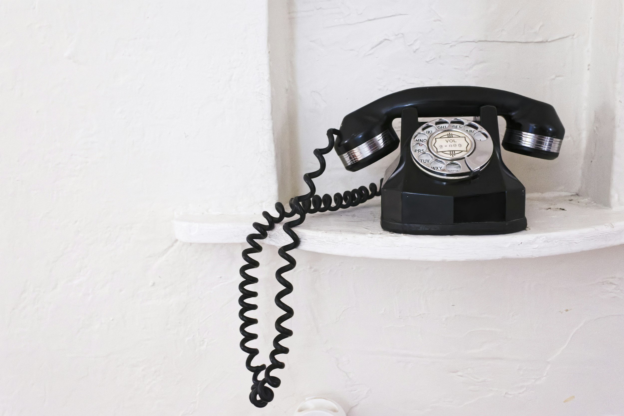 Black rotary phone on a white shelf against a textured white wall.
