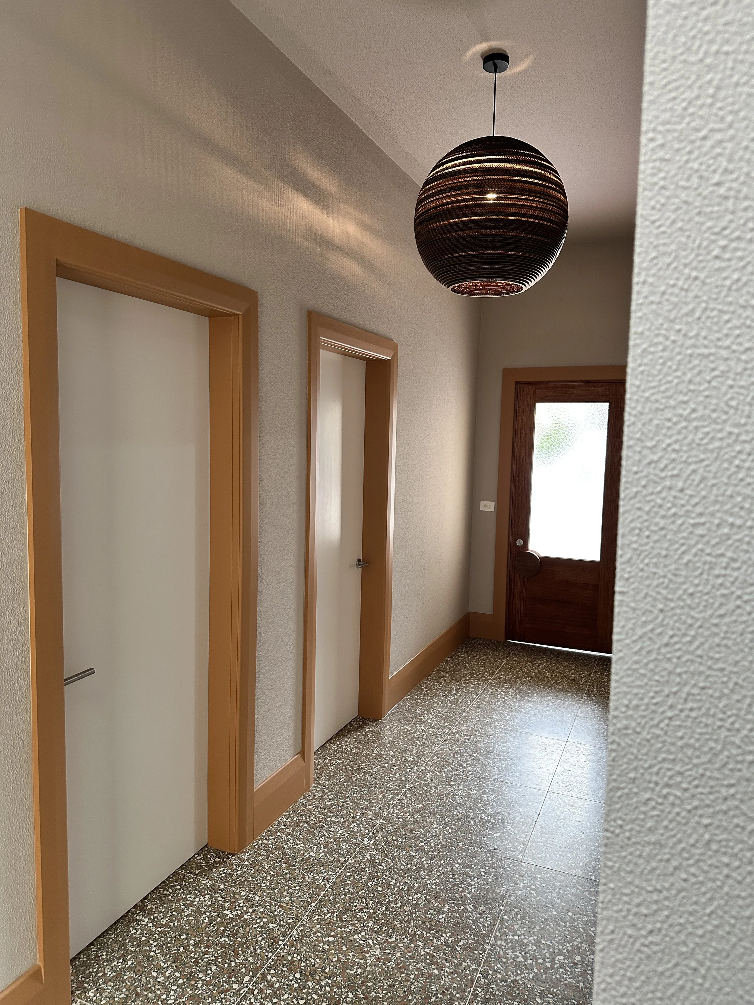 Indoor hallway with terrazzo flooring, beige textured walls, two closed white doors with wooden trim, and a wooden door with a glass panel leading outside; a round black pendant light hanging from the ceiling.