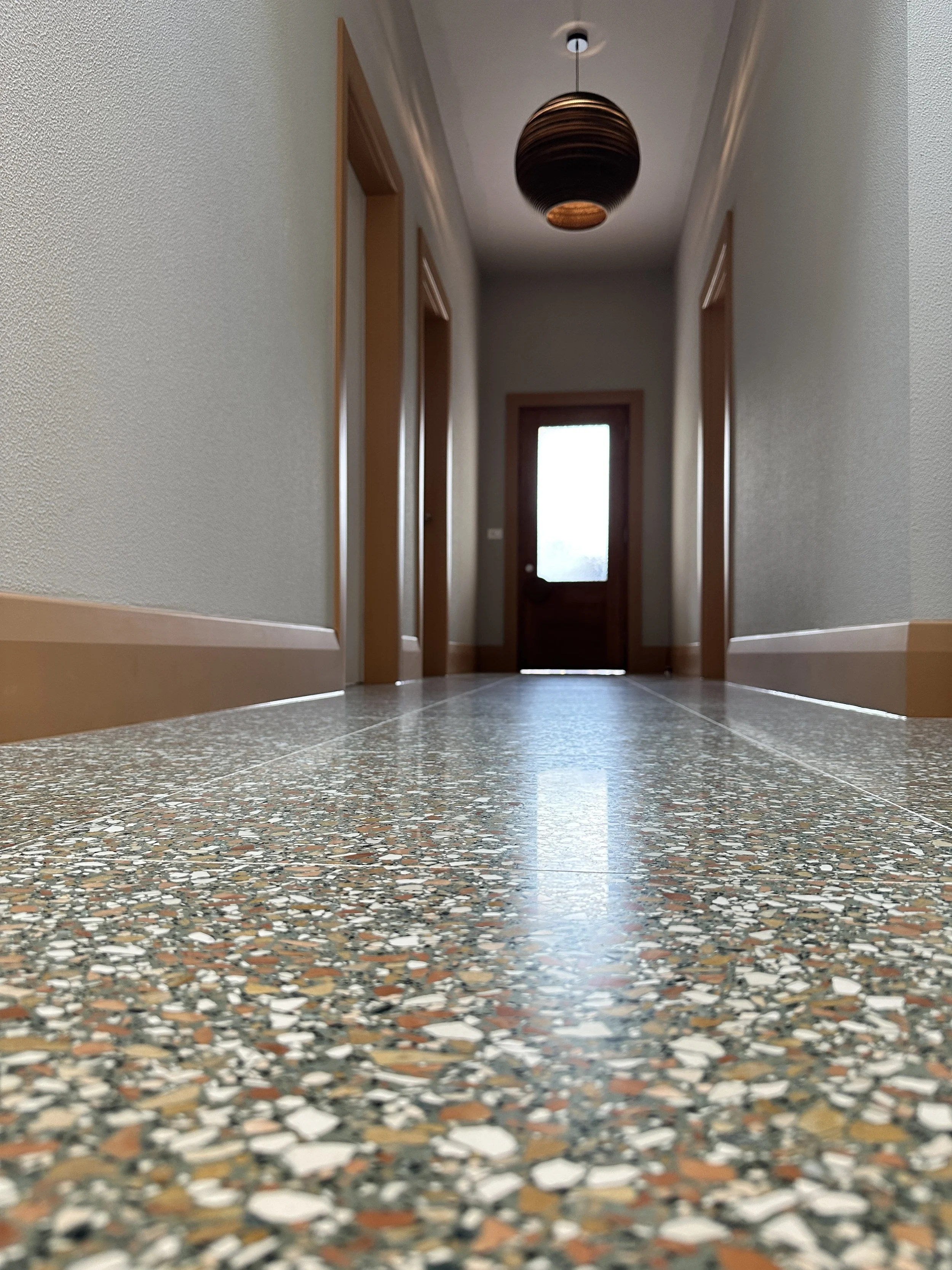 Looking down a hallway with a terrazzo floor, beige walls, wooden door frames, and a ceiling light fixture, towards a door at the end.