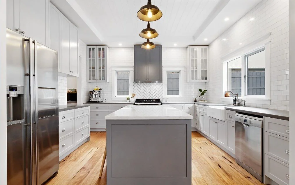 Classic white kitchen with gray cabinets, stainless steel appliances, wooden floor, central island, pendant lights, and large windows.