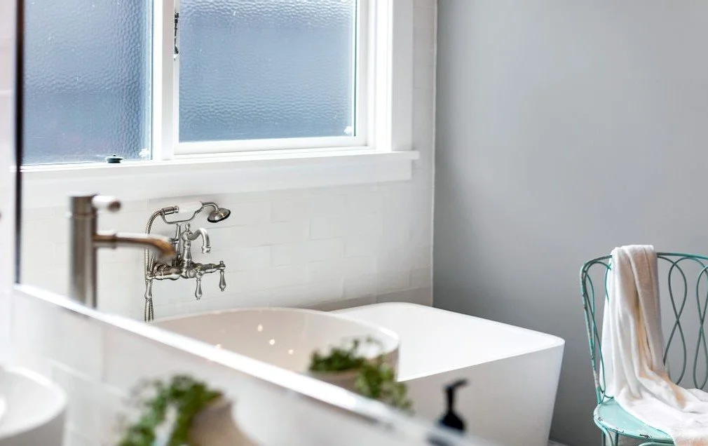 A bathroom with a white bathtub, a window with frosted glass, a wall-mounted faucet, a wire chair with a towel draped over it, and a mirror reflecting part of the room.
