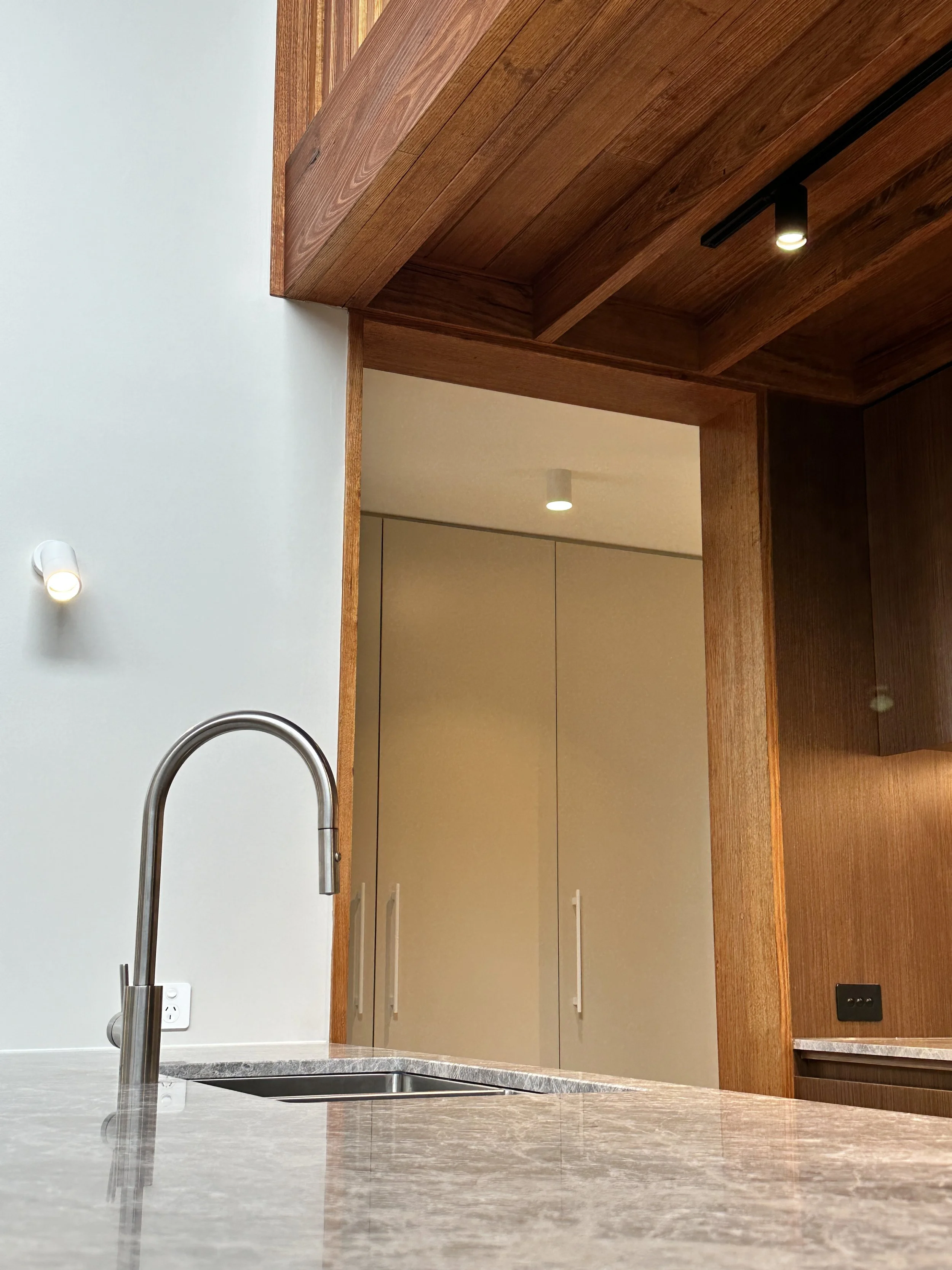 Modern kitchen with marble countertop, stainless steel faucet, white wall, built-in lighting, and beige cabinets with white handles, accented by wood paneling.