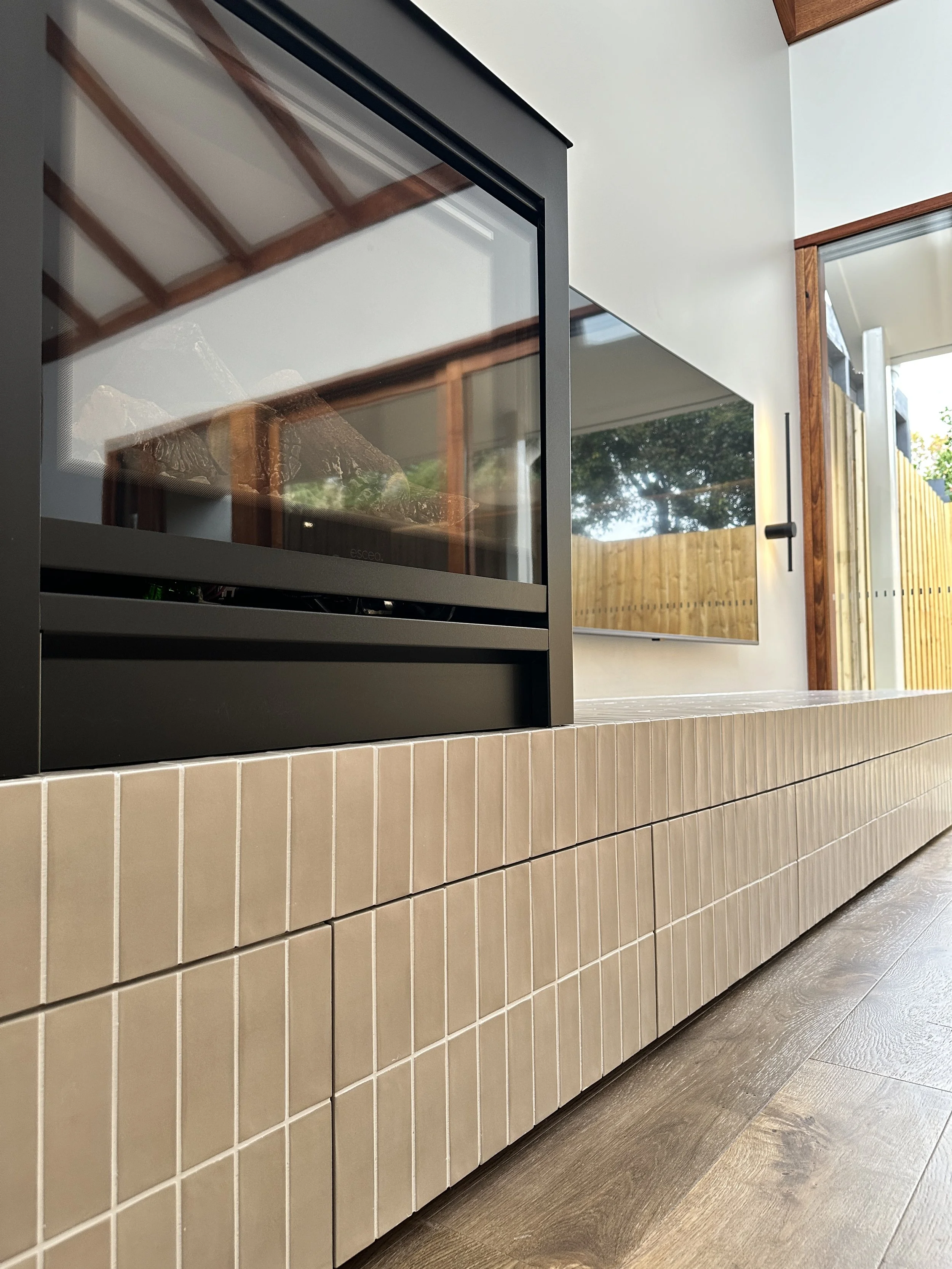 Close-up view of a modern wooden fireplace with black metal frame, beige tiled base, and a glass door, adjacent to a window with wooden trim in a contemporary interior.