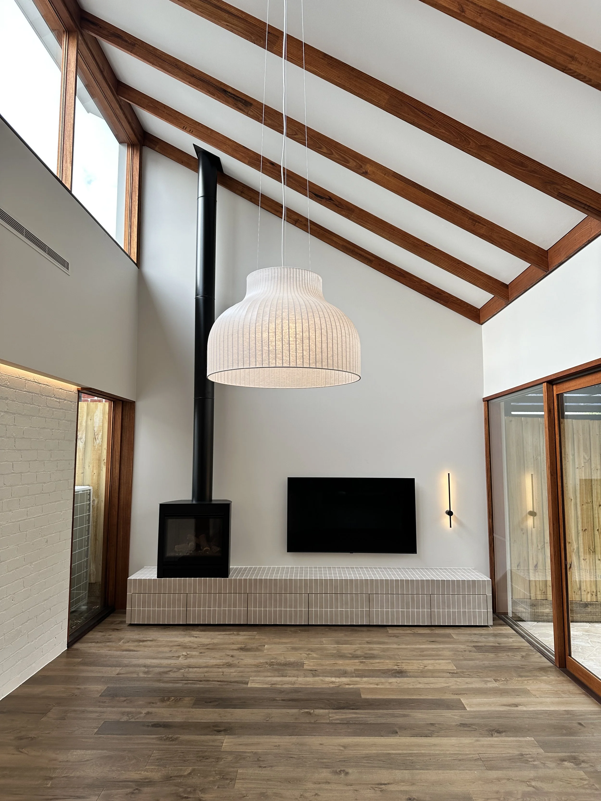 Modern living room with high vaulted ceiling, large windows, and minimalist decor. Features a wall-mounted flat-screen TV, a black fireplace with chimney, a large pendant light, and sliding glass doors leading outside.