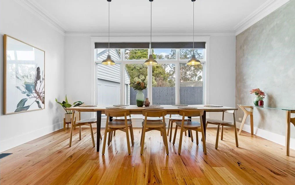 Modern dining room with a wooden table and six chairs, a large window with a view of trees, pendant lights above the table, a plant in a yellow pot on the left, artwork on the left wall, and a small table with a flower vase on the right.