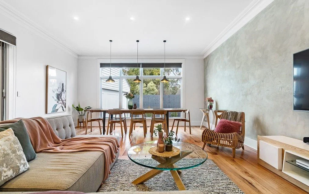 Bright living room with a gray sofa, pink blanket, and throw pillows, a glass coffee table with plants, and a greenish wall, leading to a dining area with a wooden table, chairs, and hanging pendant lights, large window with a backyard view.