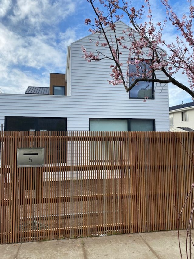 A modern house with white siding and large windows, partially obscured by a wooden fence, with a blooming pink cherry blossom tree in the foreground and a blue sky with clouds overhead.