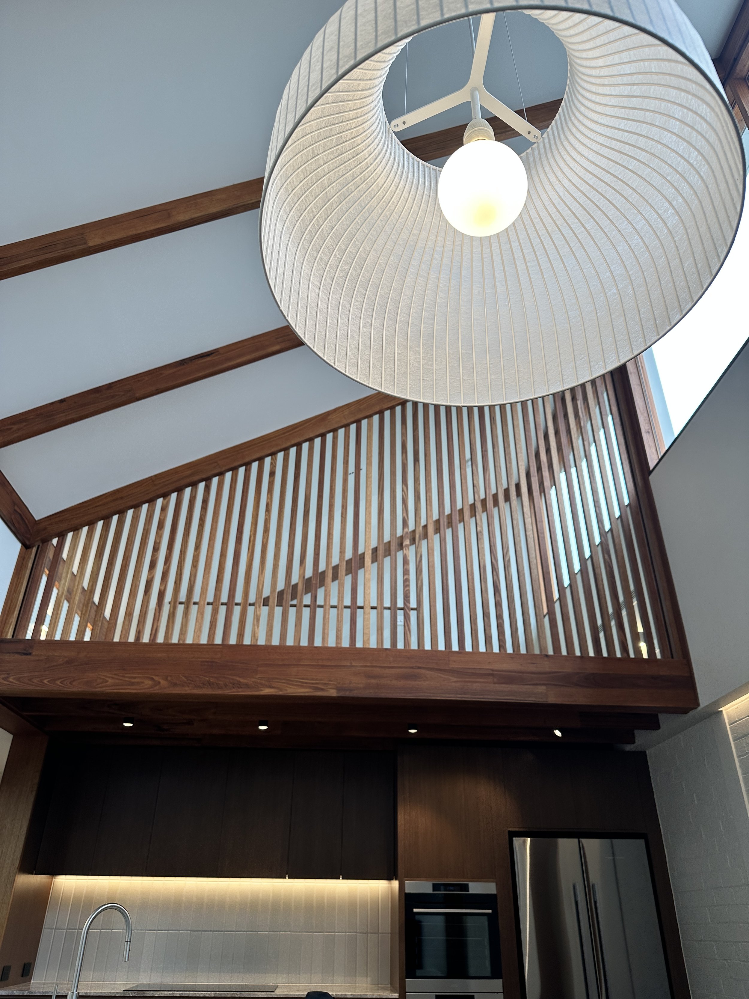 Interior view of a modern kitchen with wooden accents and a staircase with wooden railing leading to an upper level, seen from below with a large round white lampshade hanging from the ceiling.
