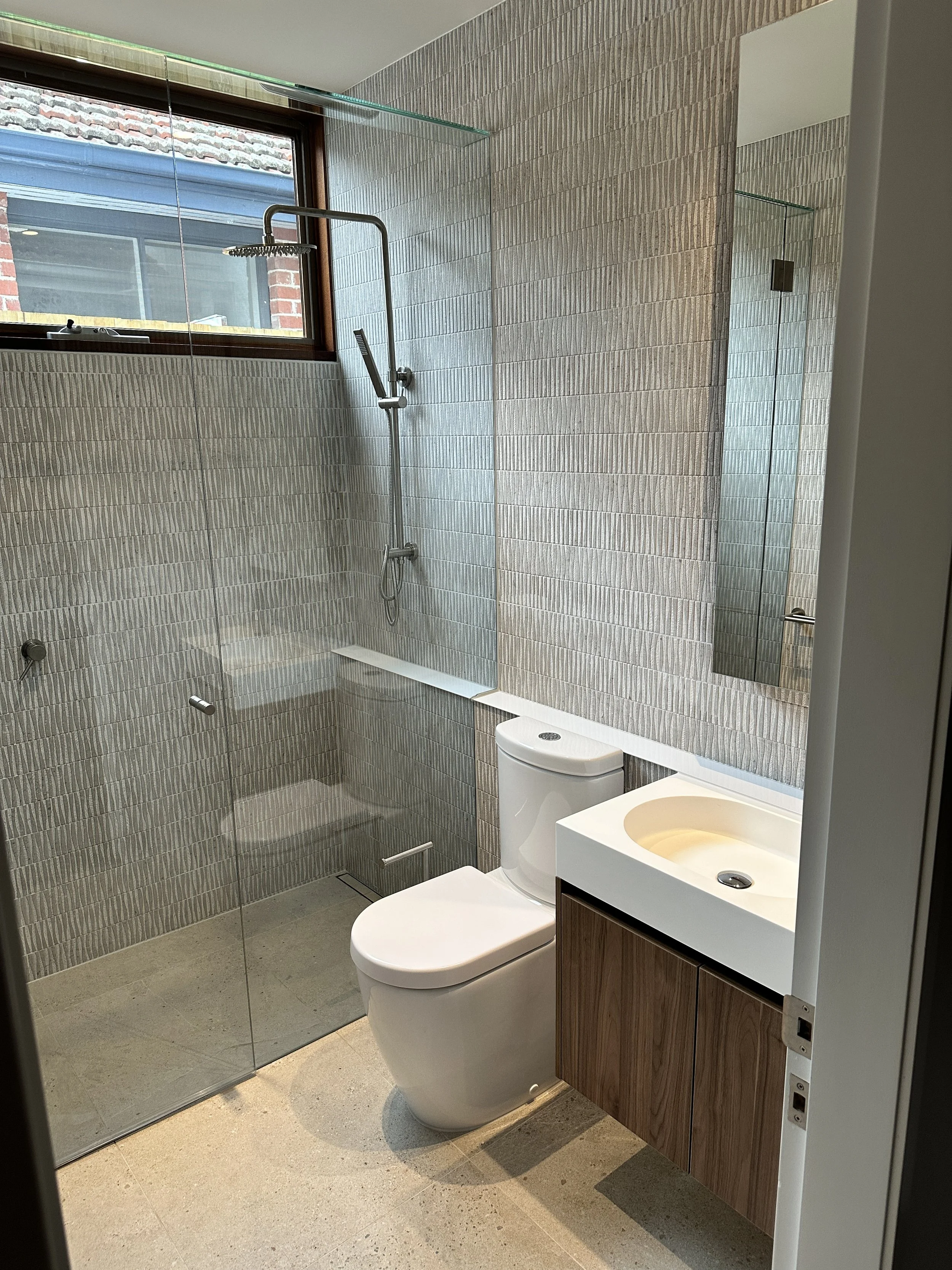 Modern bathroom with a walk-in shower featuring a glass partition, a wall-mounted showerhead, a white toilet, a small rectangular sink with a wooden cabinet, a large mirror, and a window with brick exterior visible.