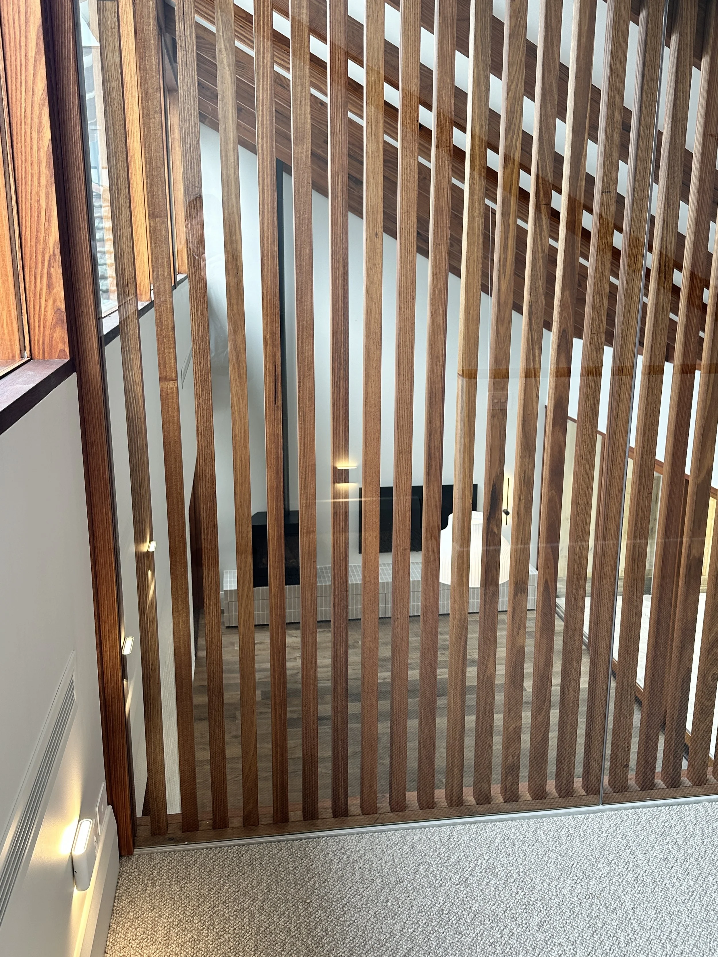 View of an interior space through wooden slats from an upper level, looking down onto a staircase landing with a television and couches visible.