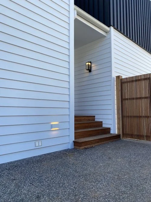 Exterior of a modern house with white siding, wooden stairs, and a black wall, with a wooden fence on the right.