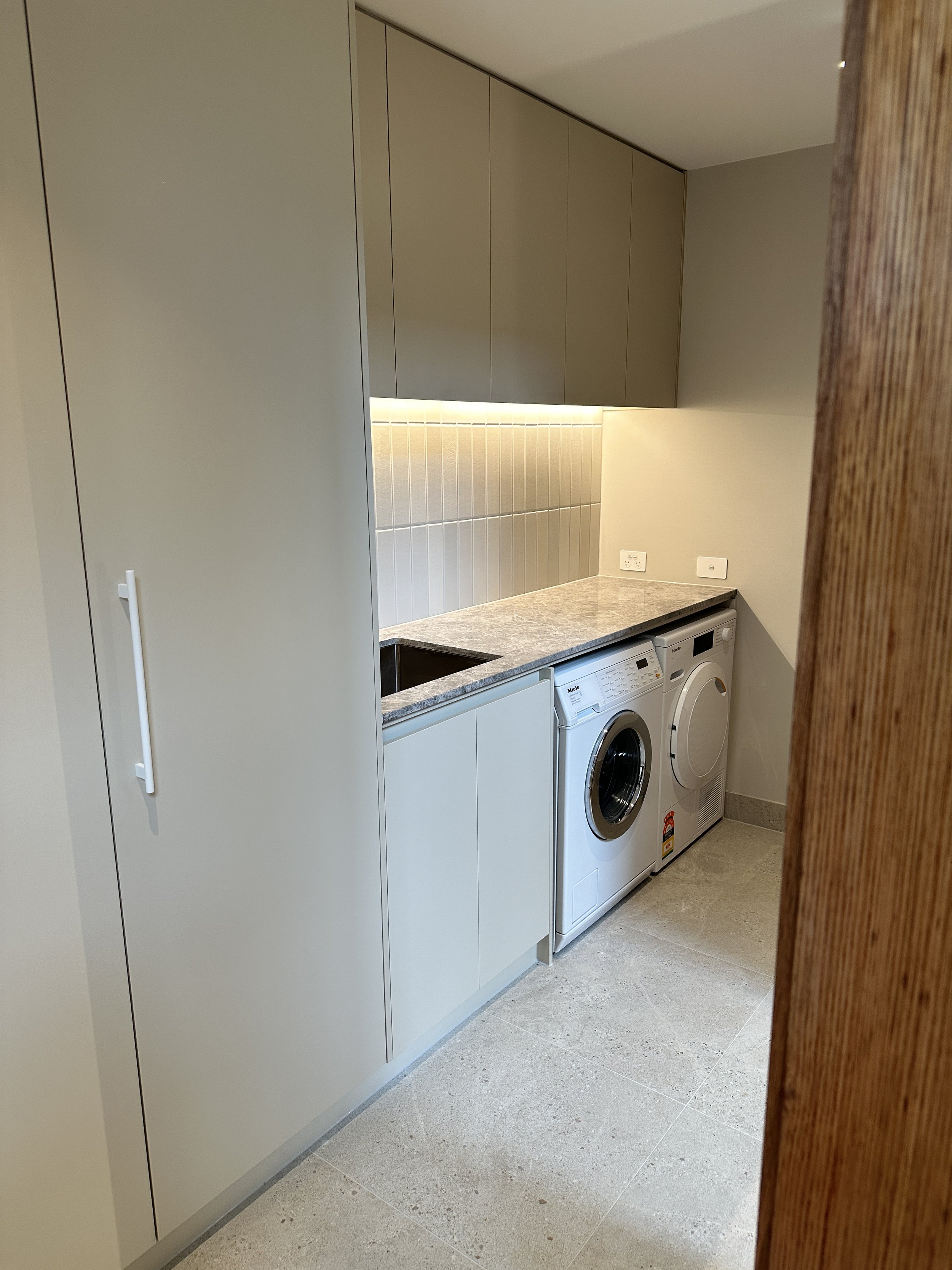 Laundry room with a washing machine and dryer beneath a gray countertop, beige cabinets above, and a tiled backsplash.