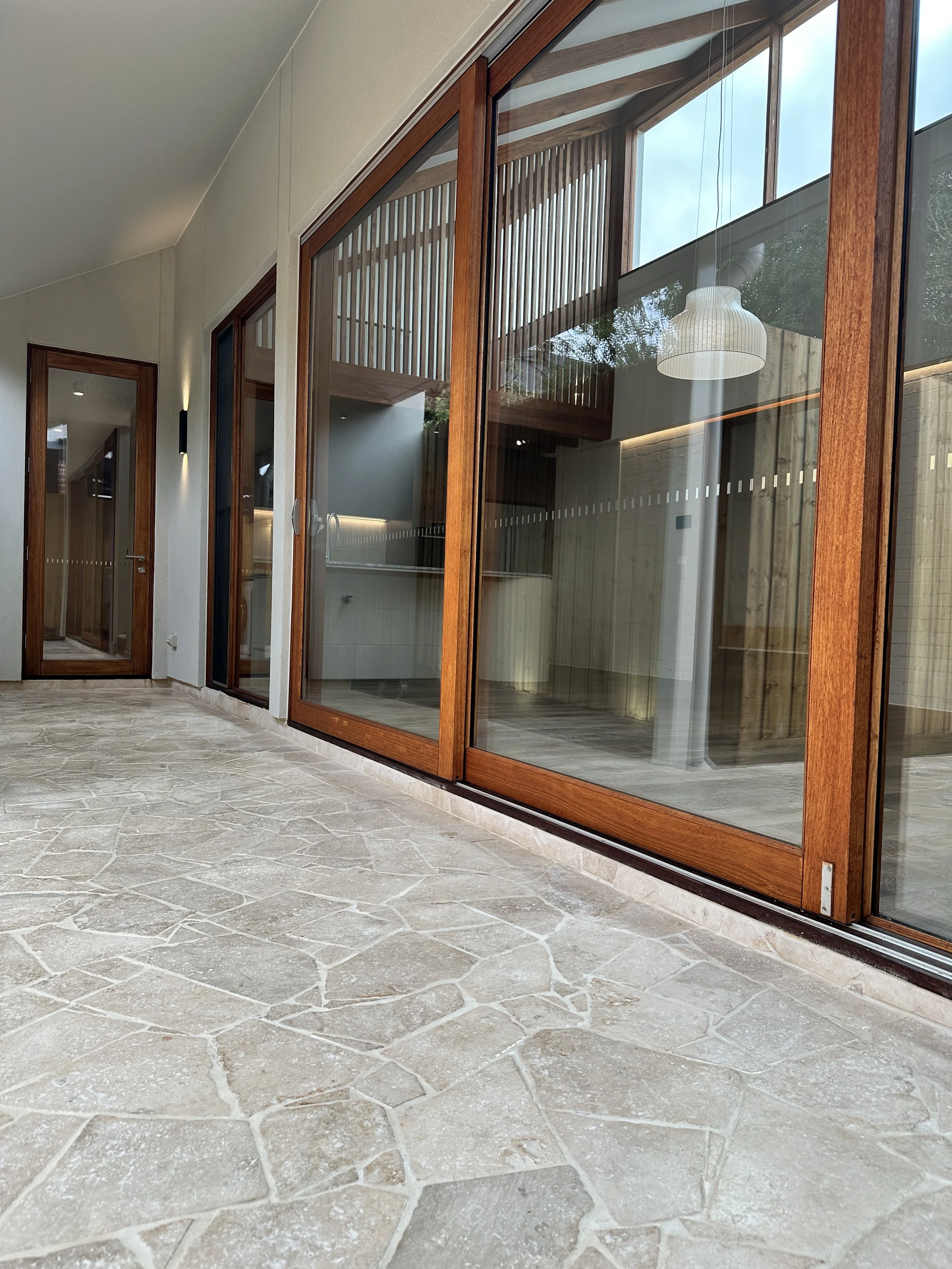 View of a modern house with large glass sliding doors with wooden frames, a stone patio, and an interior with a hanging white lamp.