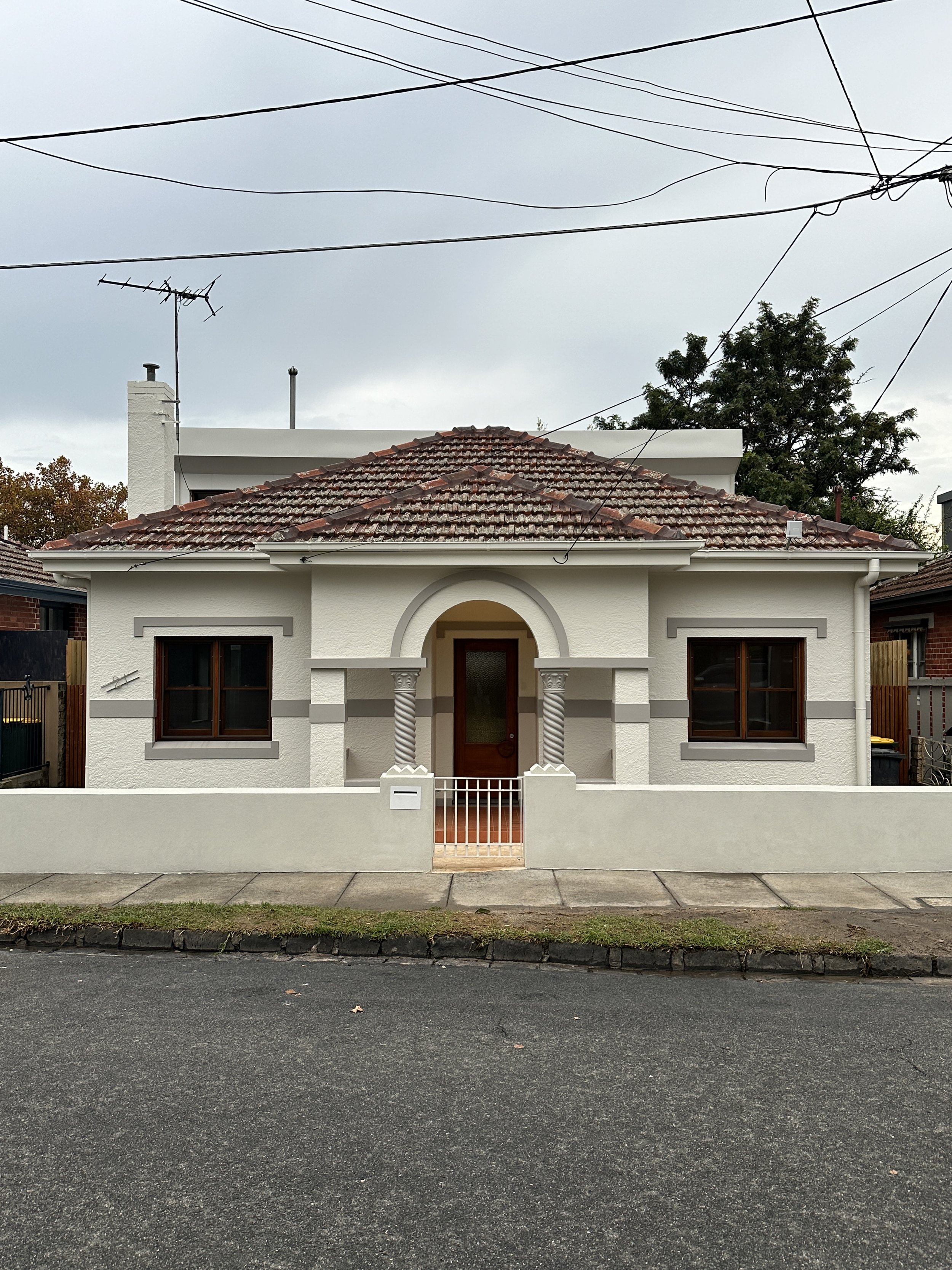 A single-story house with a red-tiled roof, white exterior walls, and two large windows with dark brown frames. The house has a central arched doorway with decorative columns and a small front gate. A sidewalk and street are in front, and there are t