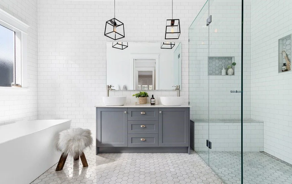Modern bathroom with white subway tile walls, a gray vanity with two vessel sinks, a large mirror, and black geometric pendant lighting. Includes a bathtub, a clear glass shower enclosure, and built-in shelves with decorative items.