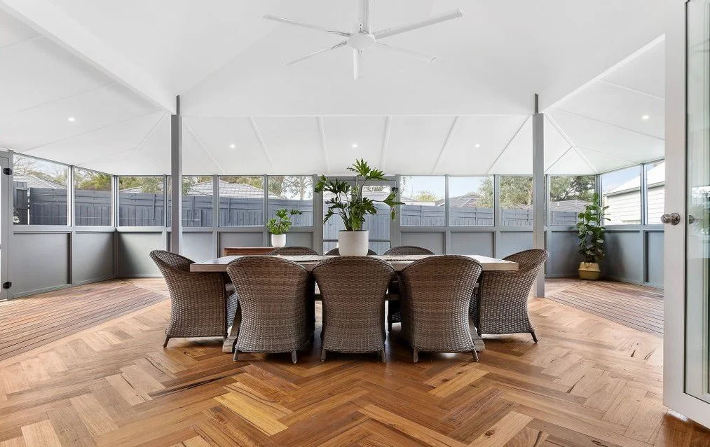 Indoor sunroom or enclosed porch with a round dining table, eight wicker chairs, potted plants, wood flooring, and large windows with a gray fence outside.