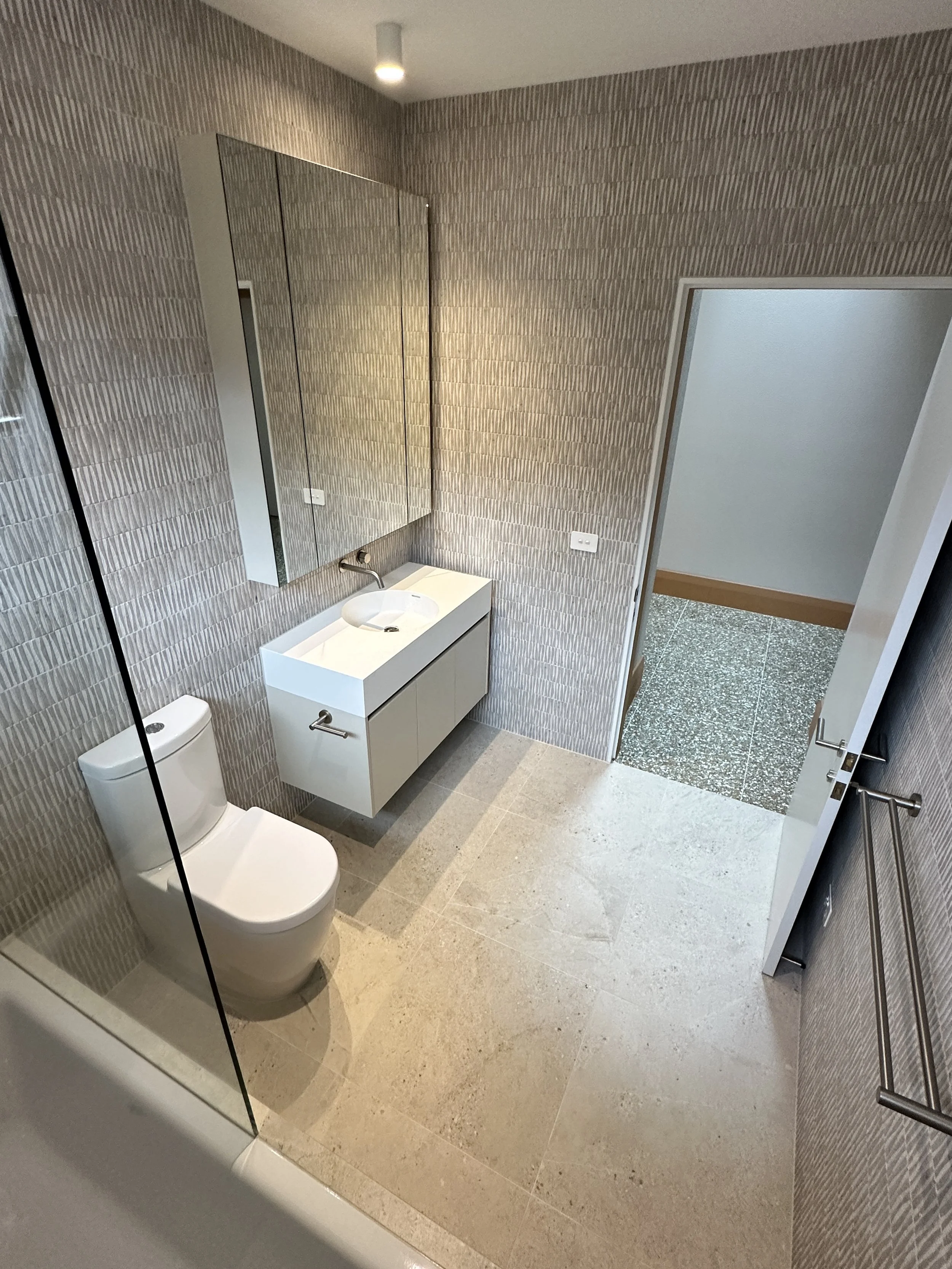 Modern bathroom with a white toilet, wall-mounted sink with cabinet, large mirror, towel bar, textured wallpaper, and an open door leading to a space with terrazzo flooring.