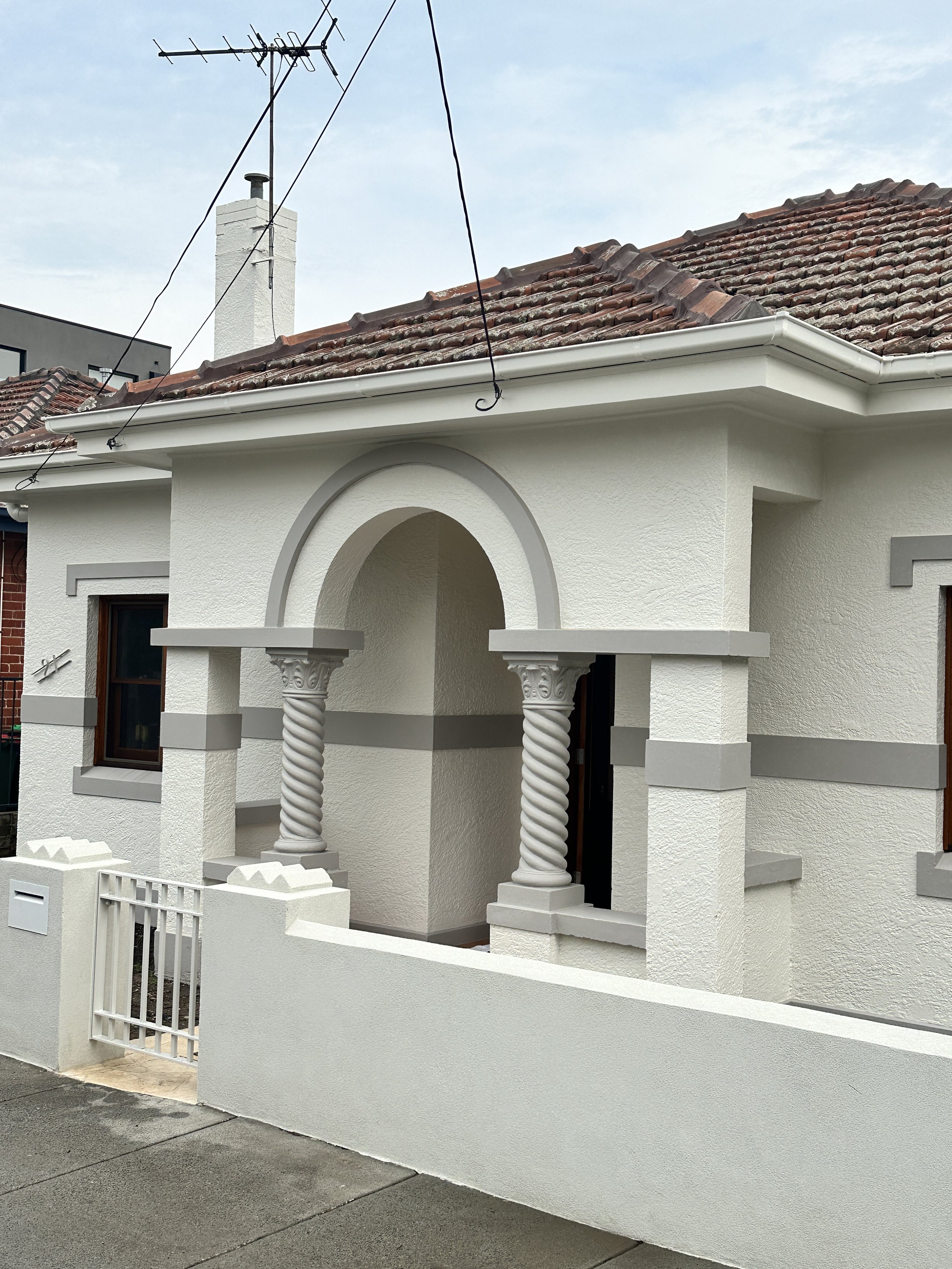 White house facade with arched porch supported by two ornate, twisted columns, a low wall, and a small gate, with a tiled roof and a chimney in the background.