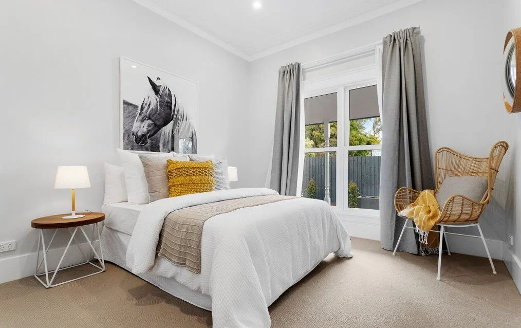 Bedroom with a white bed, framed black and white horse portrait, beige and yellow pillows, nightstand with a lamp, large window with gray curtains, rattan chair with yellow blanket, beige carpet, and white walls.