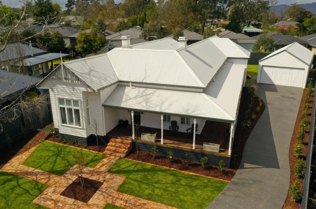An aerial view of a white house with a metal roof, a front porch with chairs, a brick walkway, a well-maintained lawn with small trees, and a driveway leading to a detached garage, in a suburban neighborhood.
