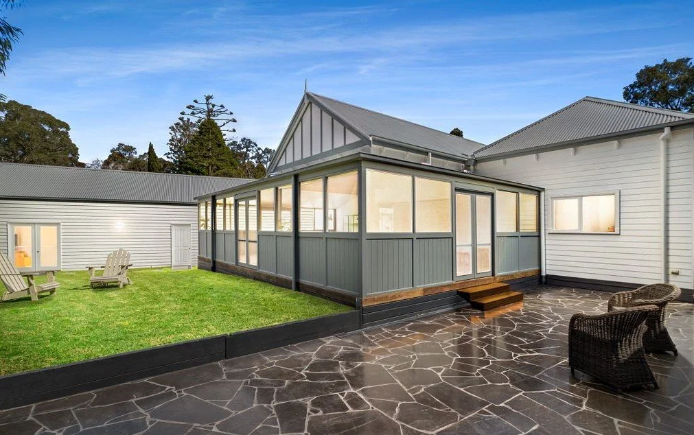 A house with a stone patio, a screened porch, and a small grassy area with Adirondack chairs. The house has white siding and metal roofs, and the background shows trees and a blue sky.