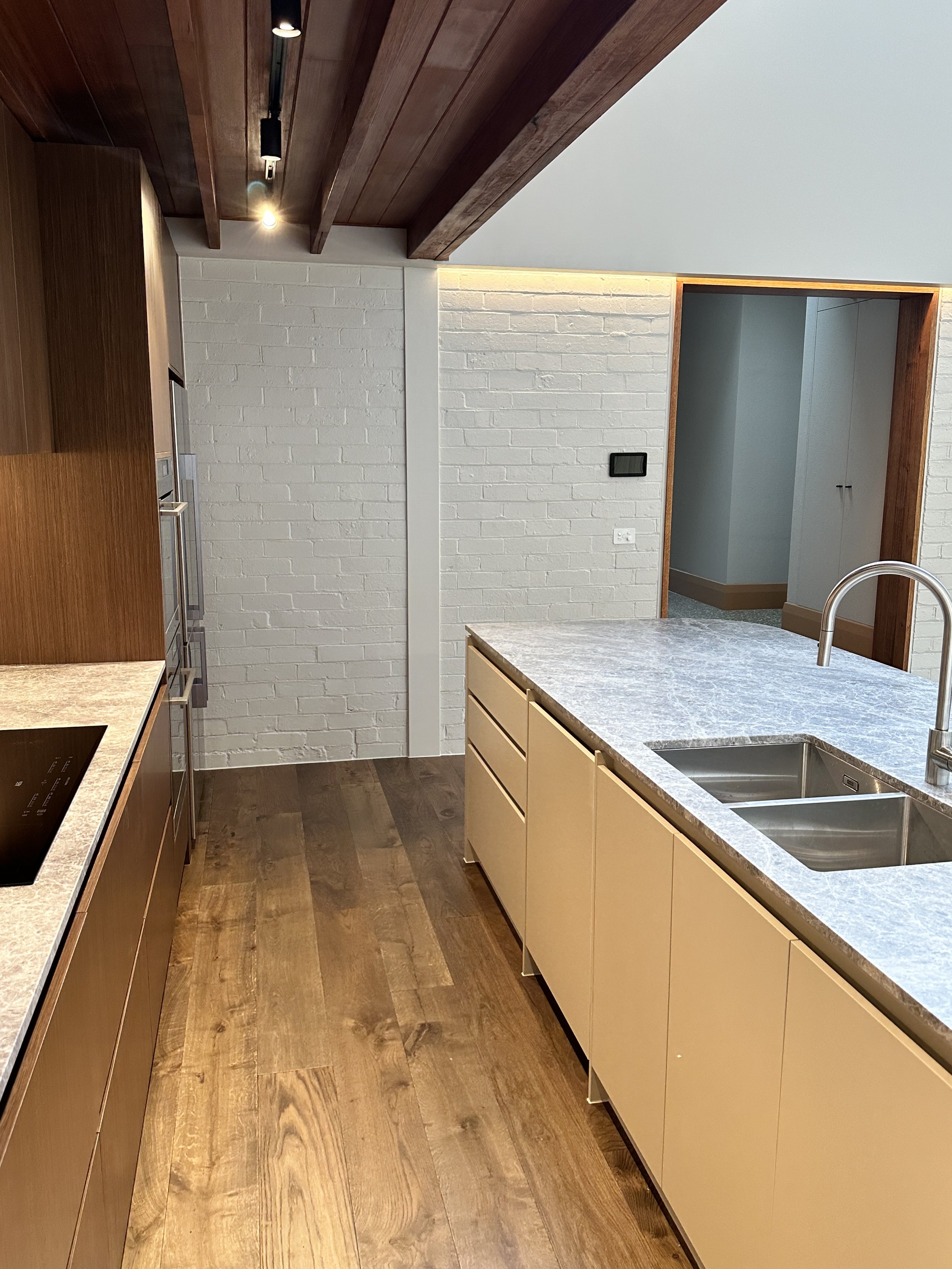 Modern kitchen with white brick wall, wooden ceiling beams, beige cabinets, gray marble countertop, stainless steel sink, and wooden flooring.