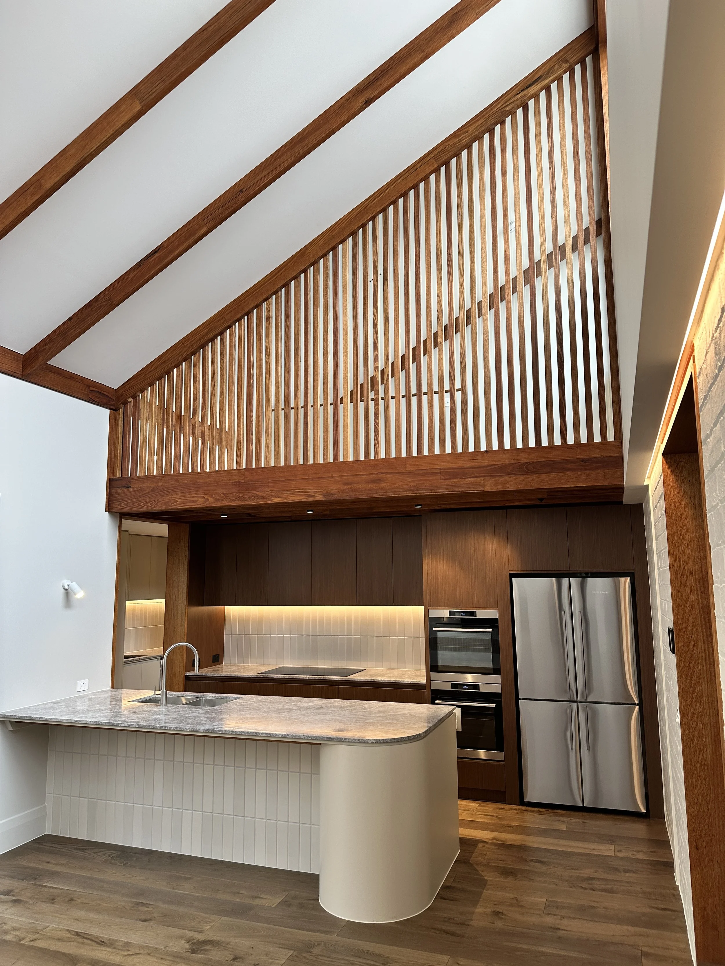 Modern kitchen with a curved island, wooden cabinetry, stainless steel appliances, upper wooden slat railing, and a white ceiling with exposed wooden beams.