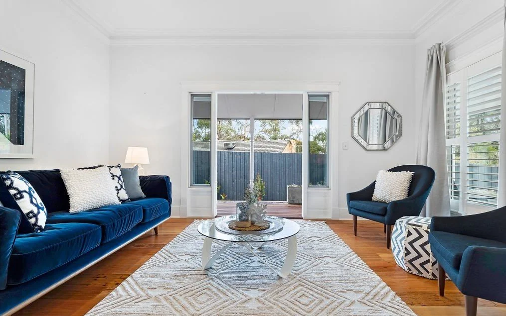 Living room with a navy sofa, two navy armchairs, a glass coffee table, white area rug, large windows, and a sliding glass door leading to an outdoor patio.