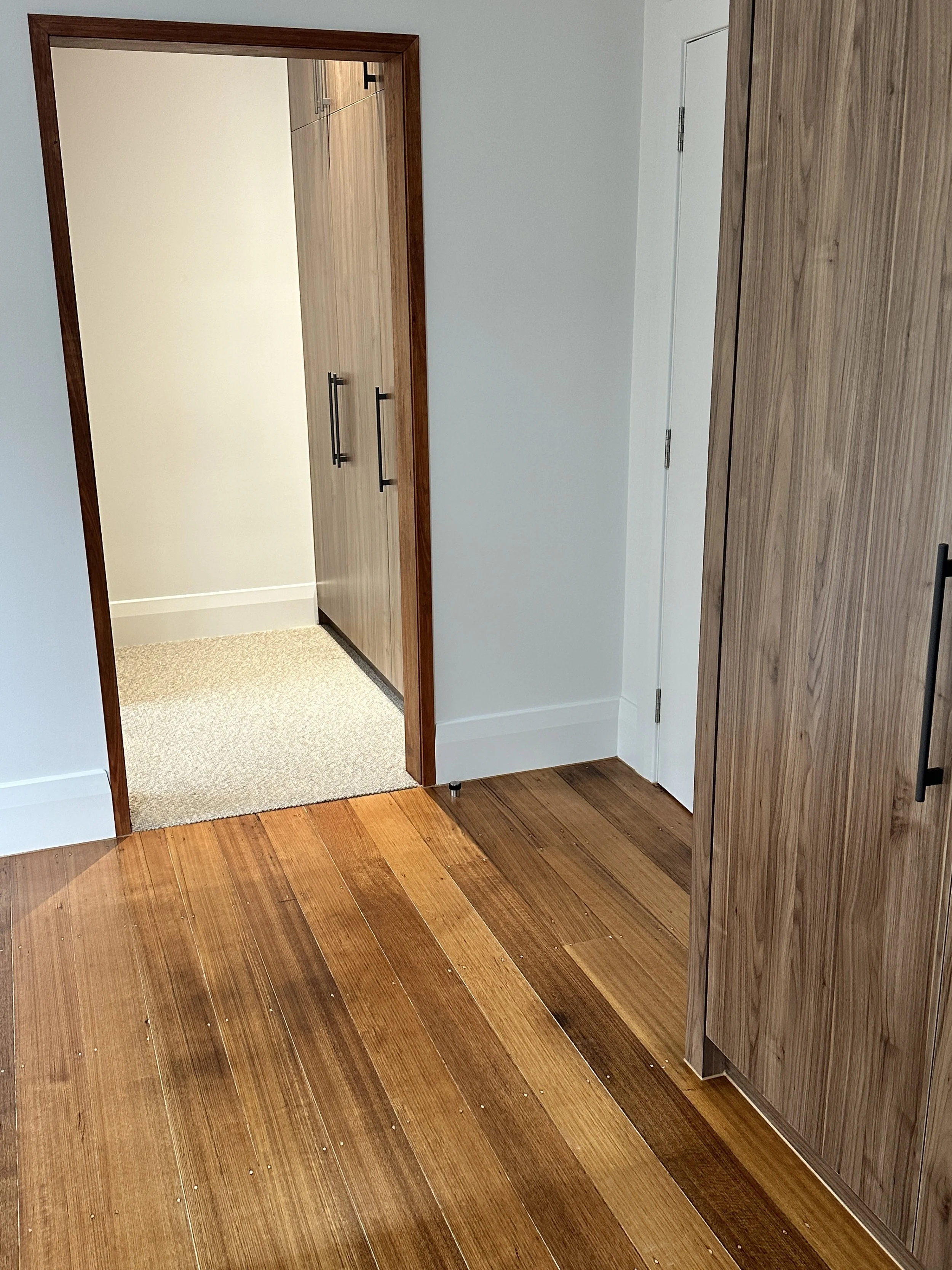 A room with wooden flooring leading to a doorway with a wooden trim and a small hallway with beige carpet. There are white walls, a white door, and wooden cabinets on the right.