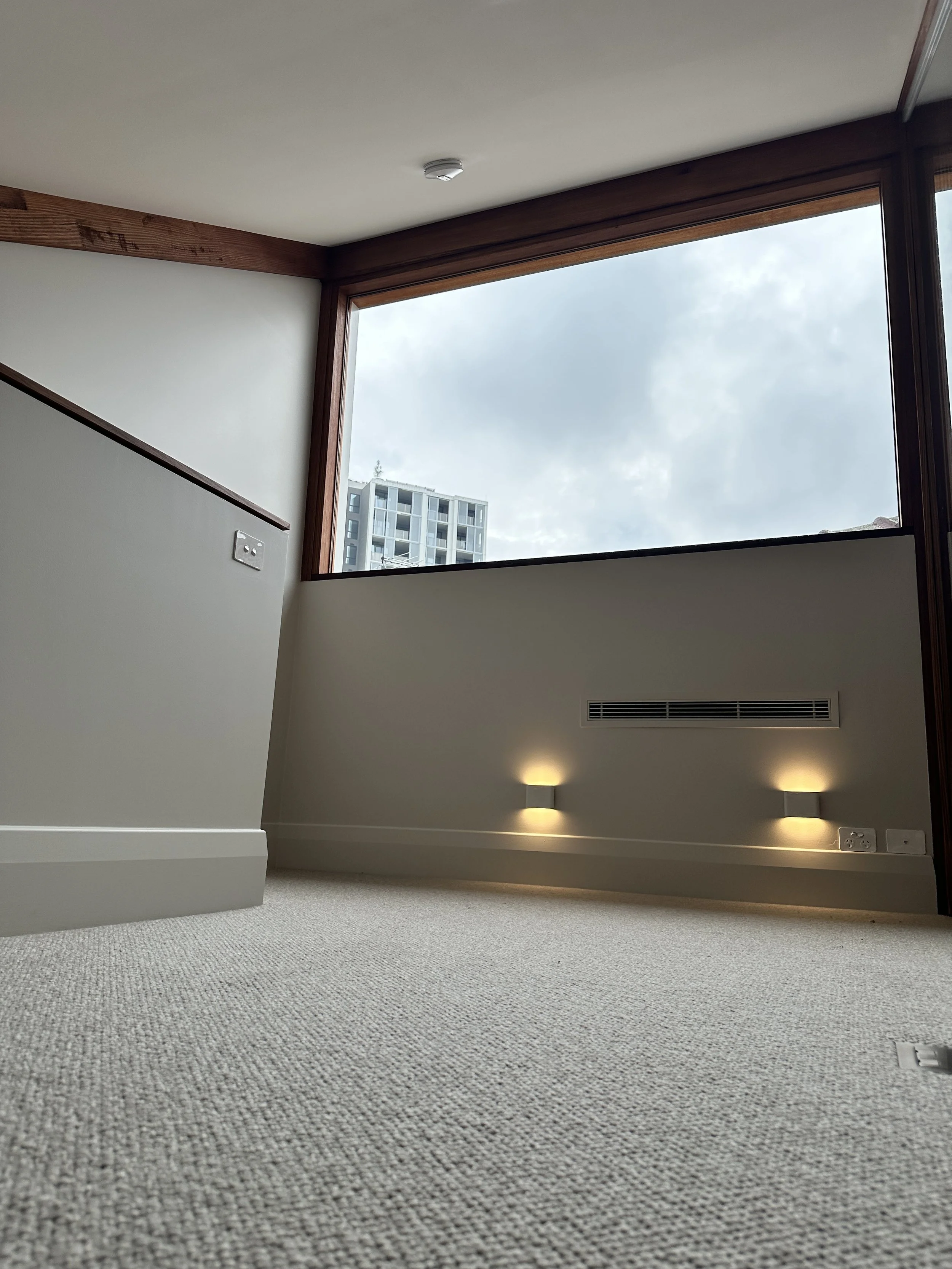 Empty room with beige carpet, large window showing an outside building and cloudy sky, wall lights illuminating the wall, and wooden framing around the window.